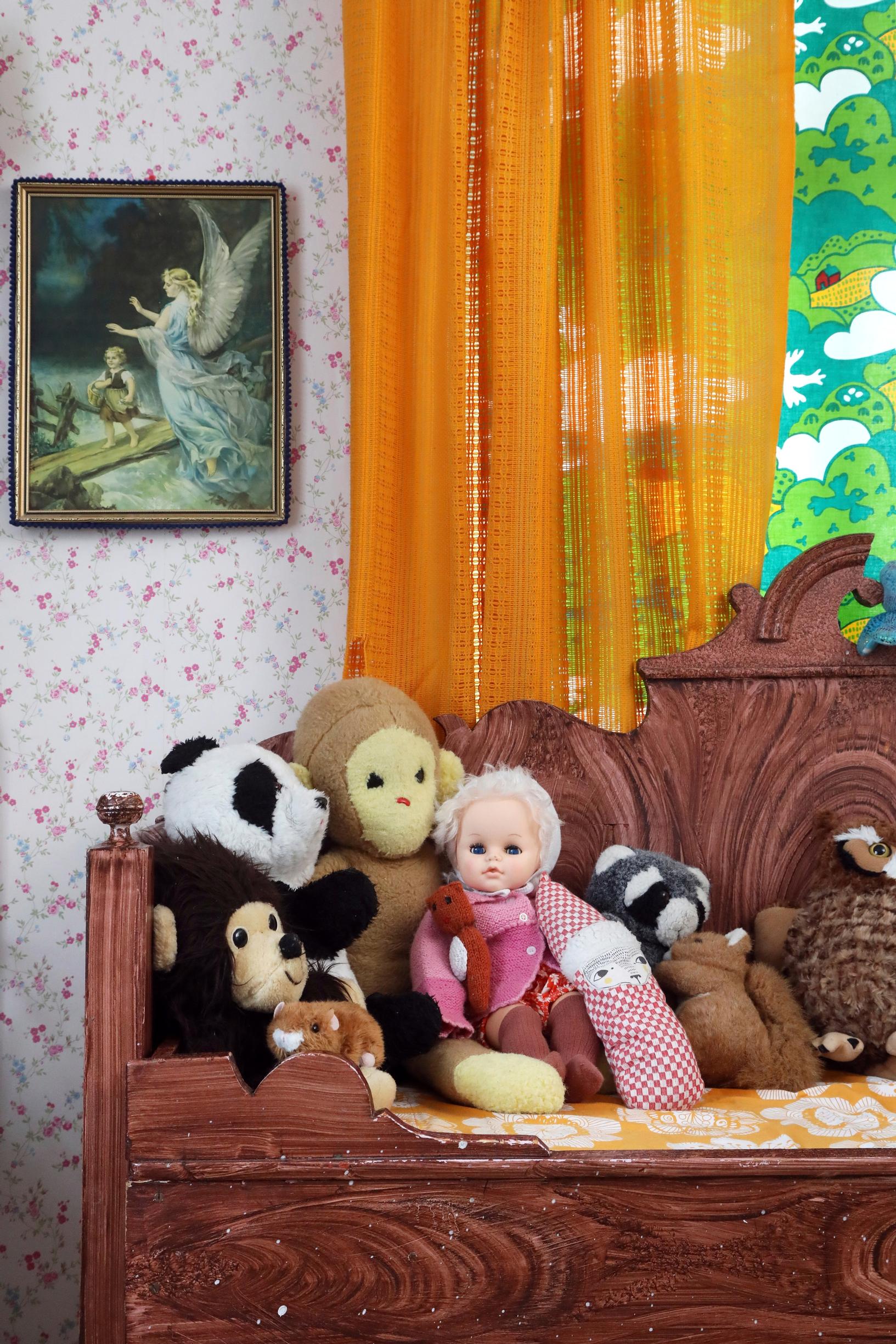 A doll collection on the children’s room wooden sofa, backed by a traditional Guardian Angel image.