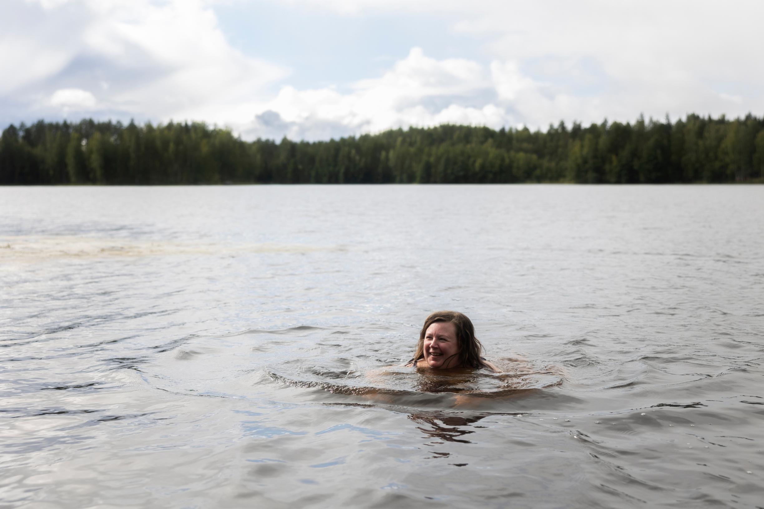 A smiling woman swimming in a lake, with a forest on the opposite shore.