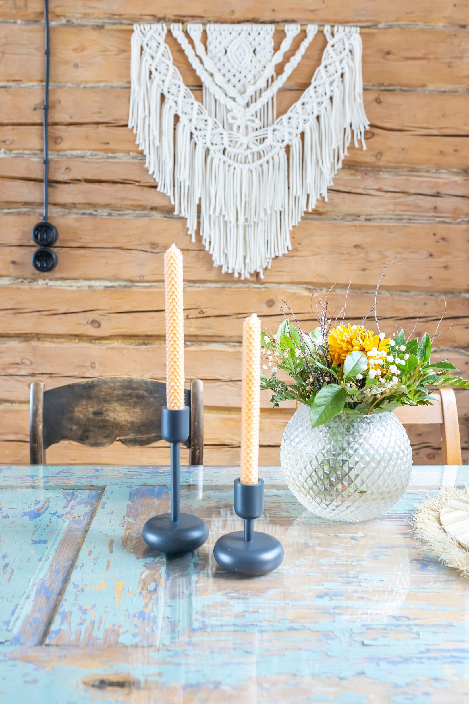 On a log wall, a macramé piece; a table with a glass top, flowers in a vase, two candleholders with candles, and chairs