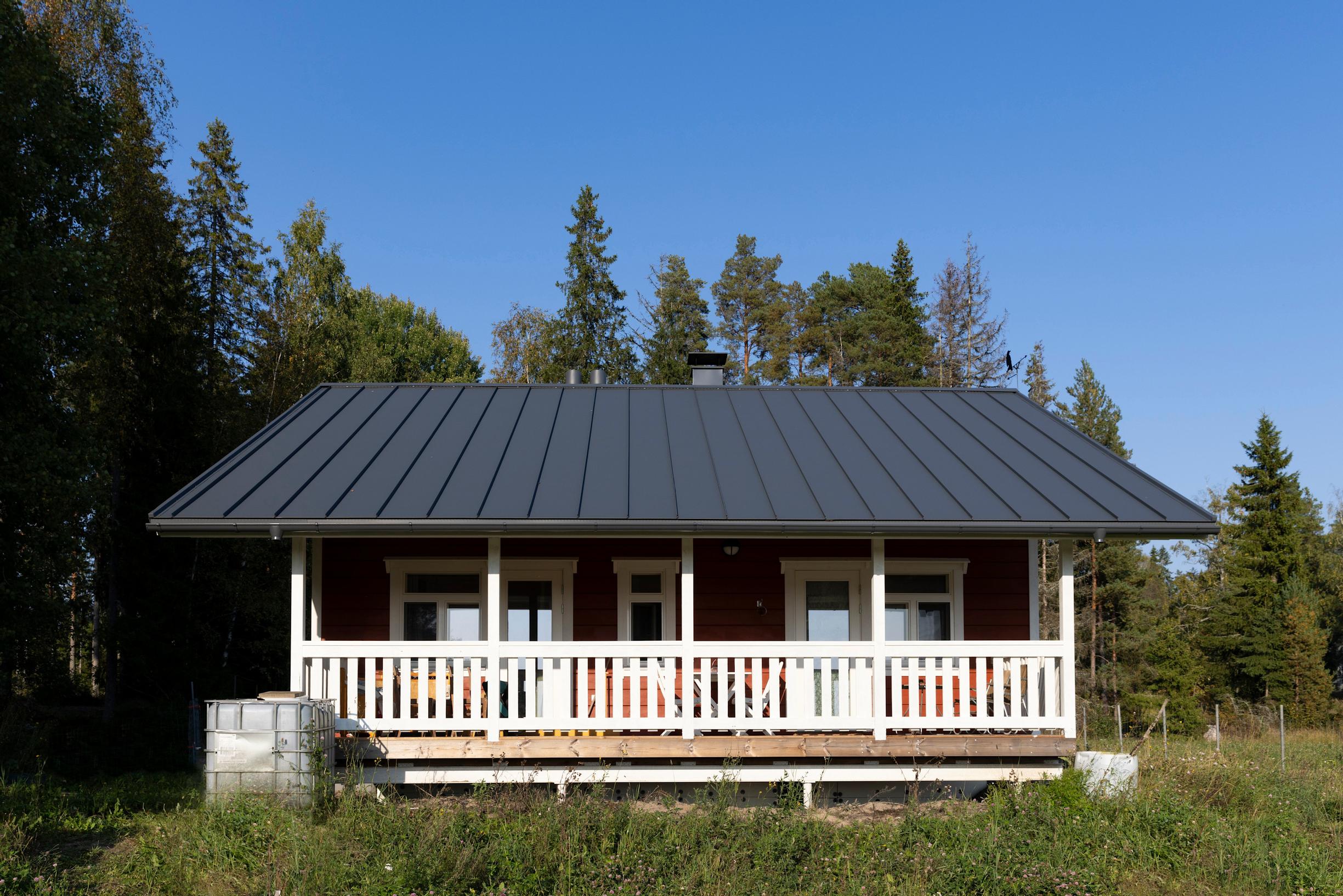 A gray-roofed house at the edge of a field and forest, with white wooden railings on the terrace.