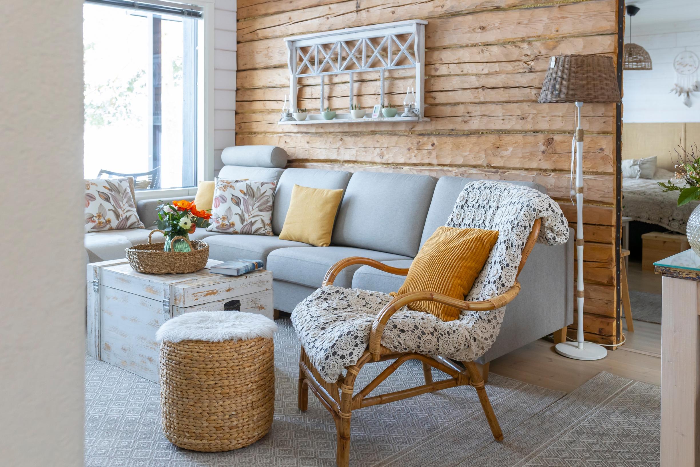 A light-colored sofa with colorful cushions, a rattan armchair, a footstool, and a chest; a log wall and view into the bedroom