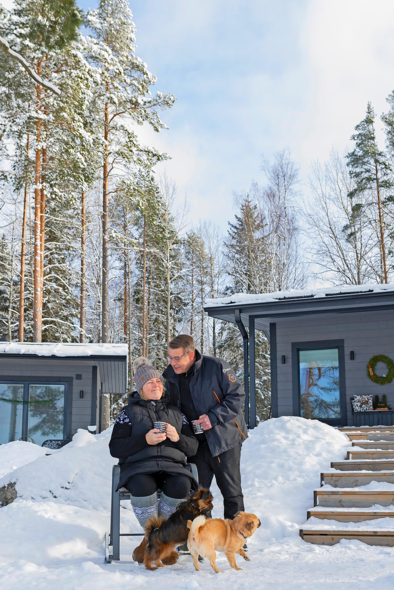 Mari and Kari sitting in the cottage garden with their two dogs, two buildings in the background, snow, and forest