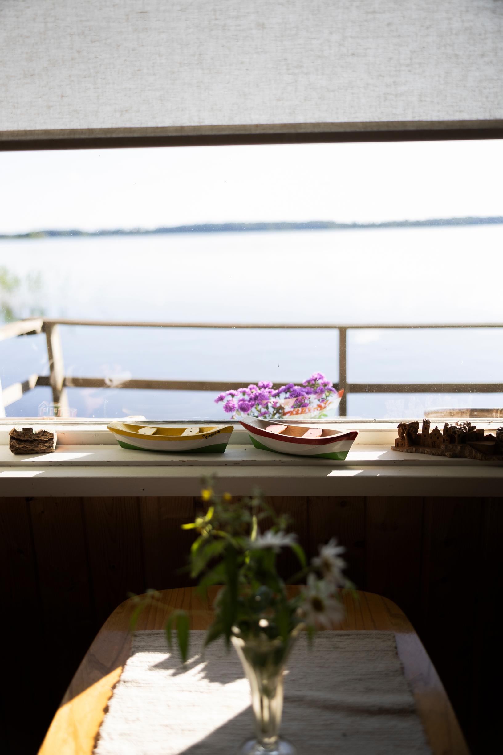 Wooden ships on the windowsill, a flower bouquet on the table in front, view of the lake and veranda railing in the background