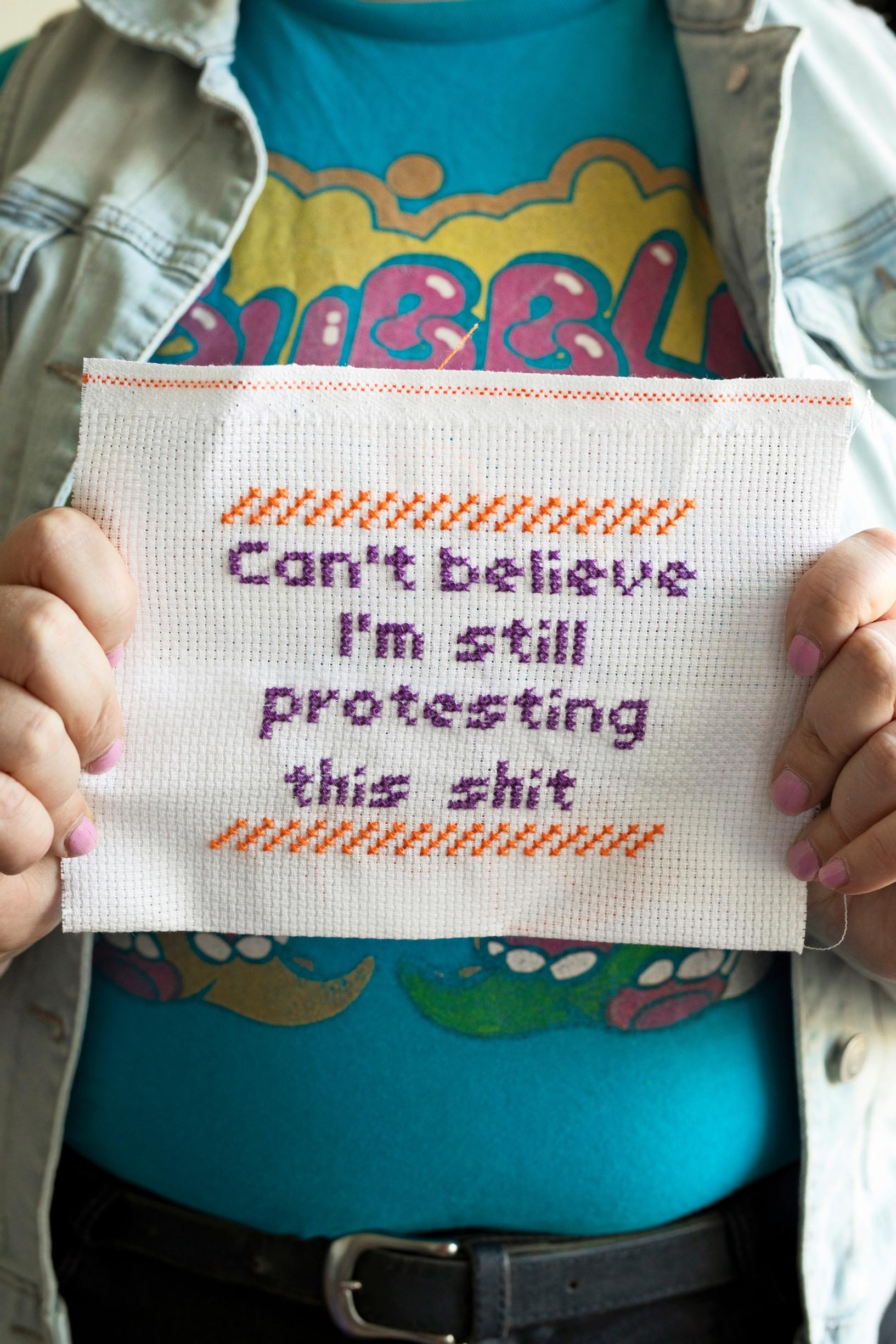 A woman holds a cross-stitch piece that reads ‘Can’t believe I’m still protesting this shit’