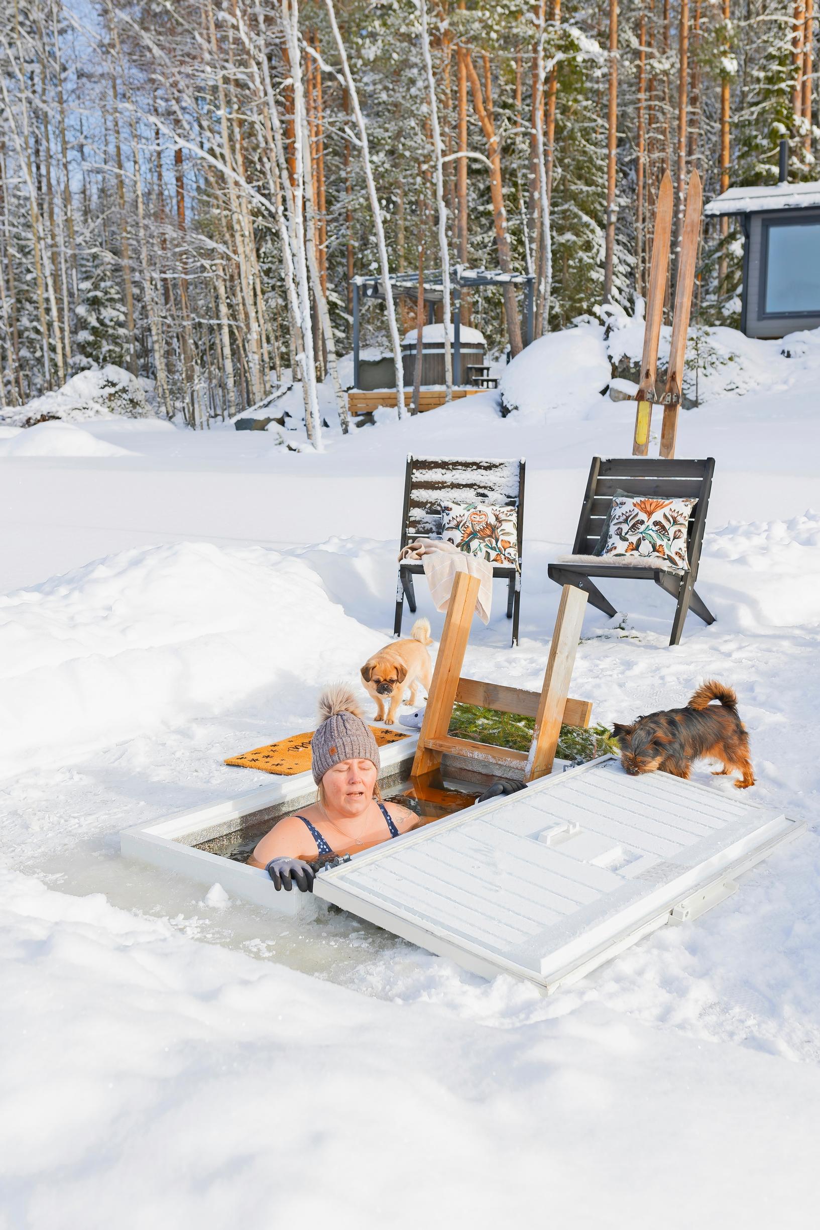 Mari in the ice hole, with the freezer chest lid and a dog nearby, ladder, two sun loungers, hot tub, and building in the background