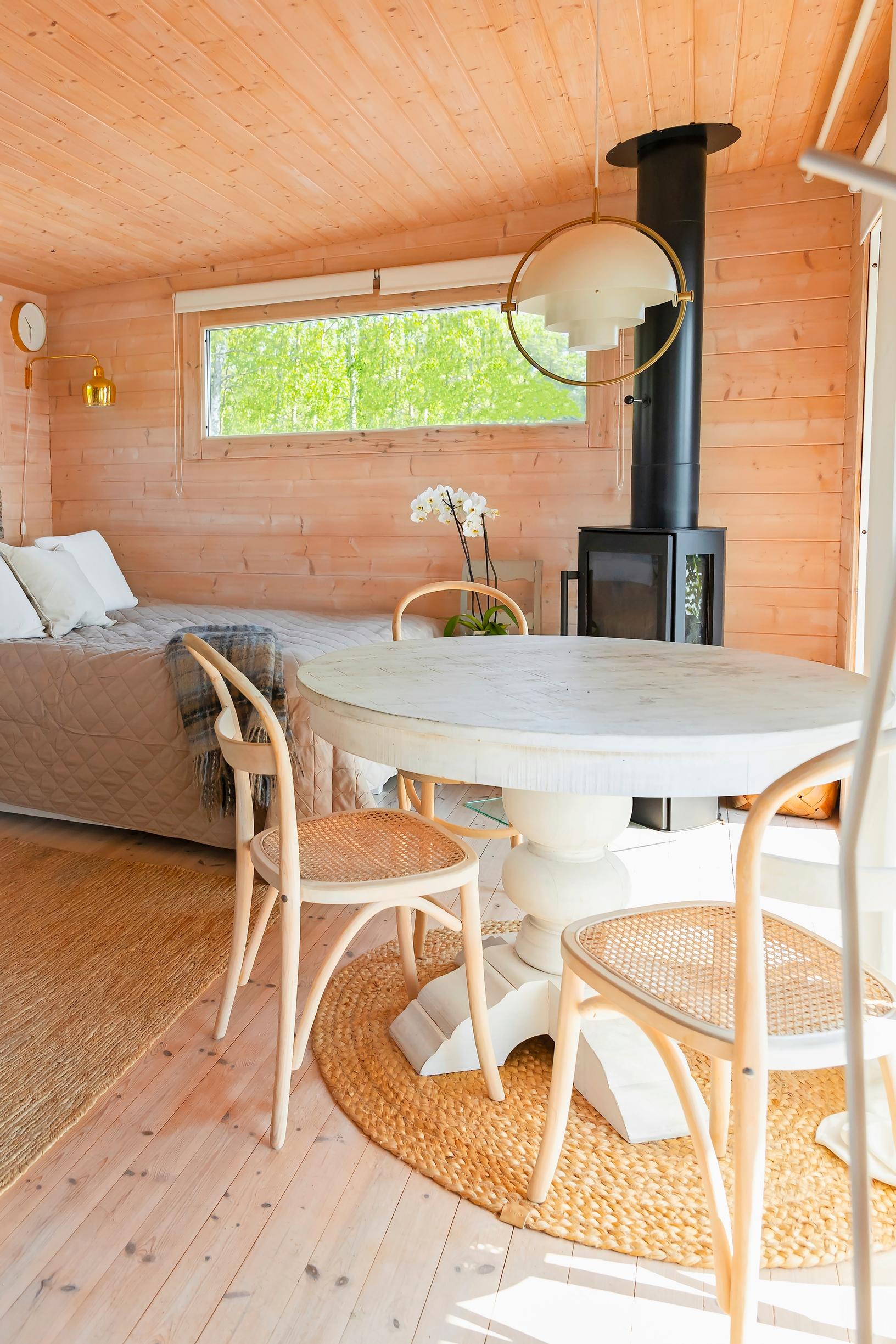 a round table and brown chairs, a black wood stove, and a bed in the cabin