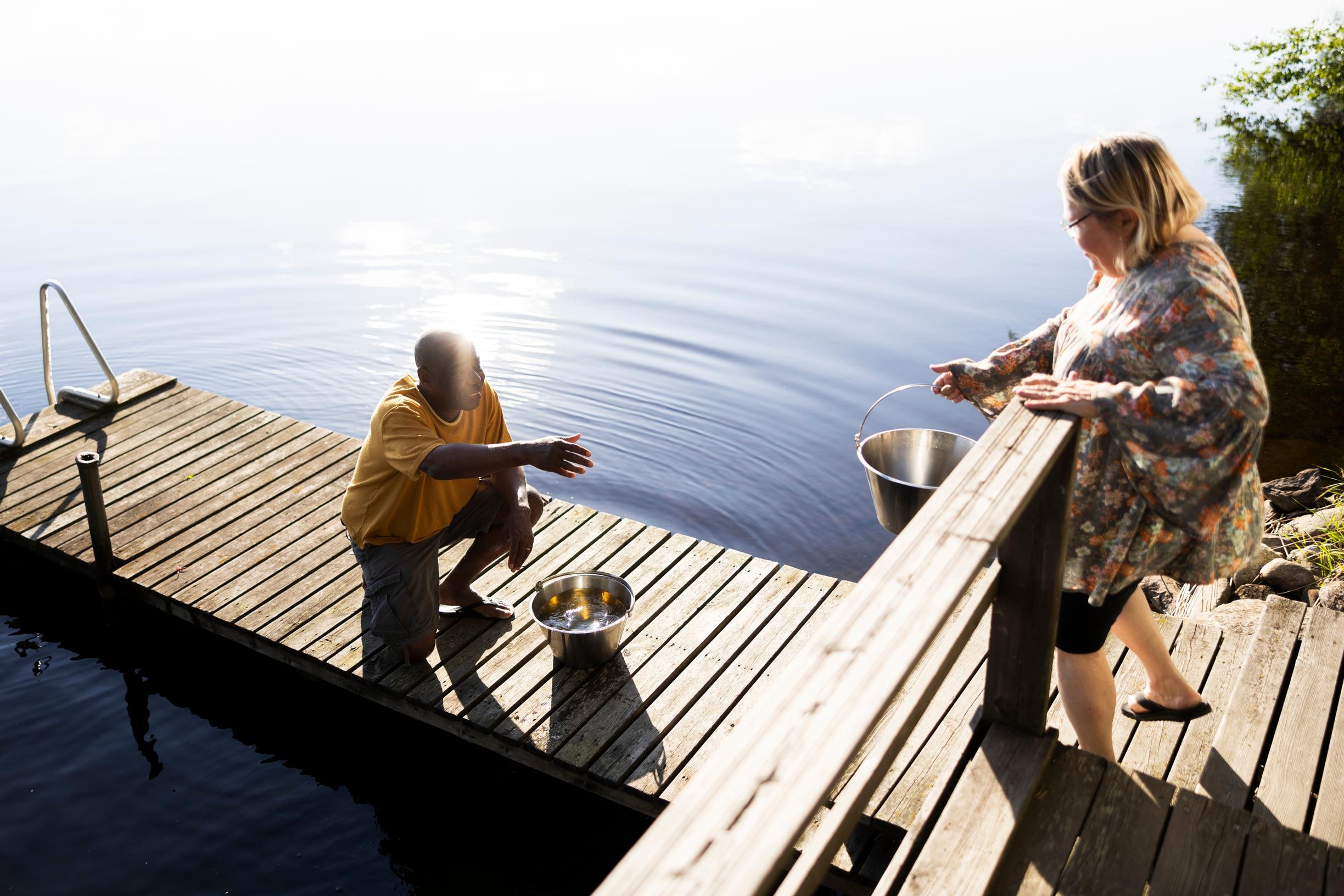 Edson crouching on the dock, receiving a metal bucket handed by Helena