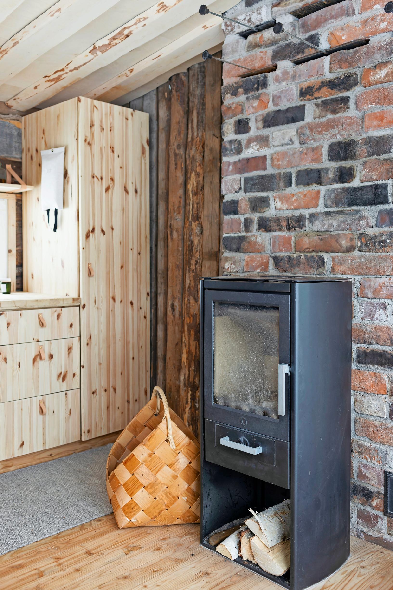 A wood-burning stove in front of a vintage brick wall, accompanied by a woven wooden basket and a pine cabinet.