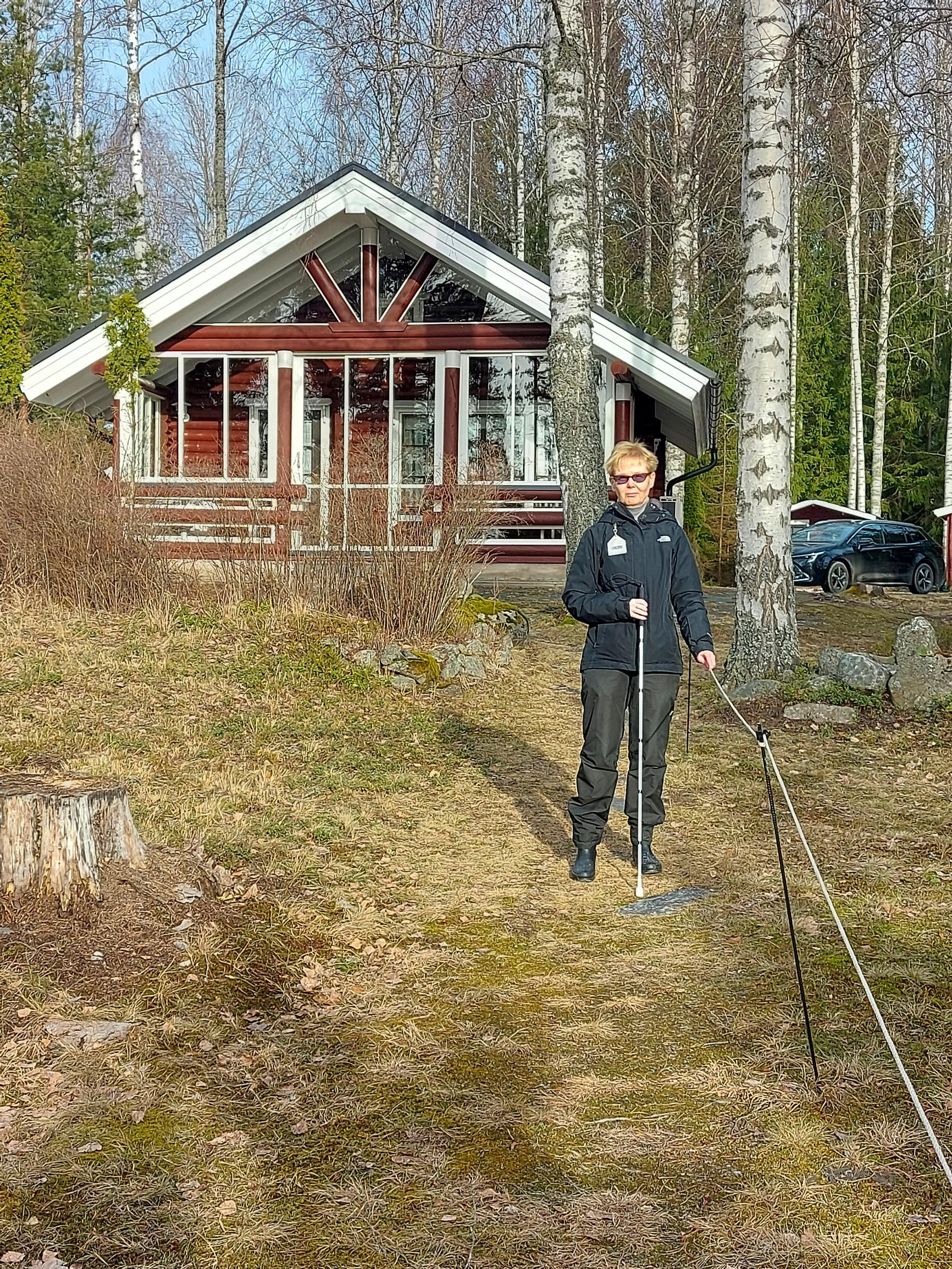 Merja Heikkonen holds a guide rope in one hand and a cane in the other, with a red summer cottage in the background