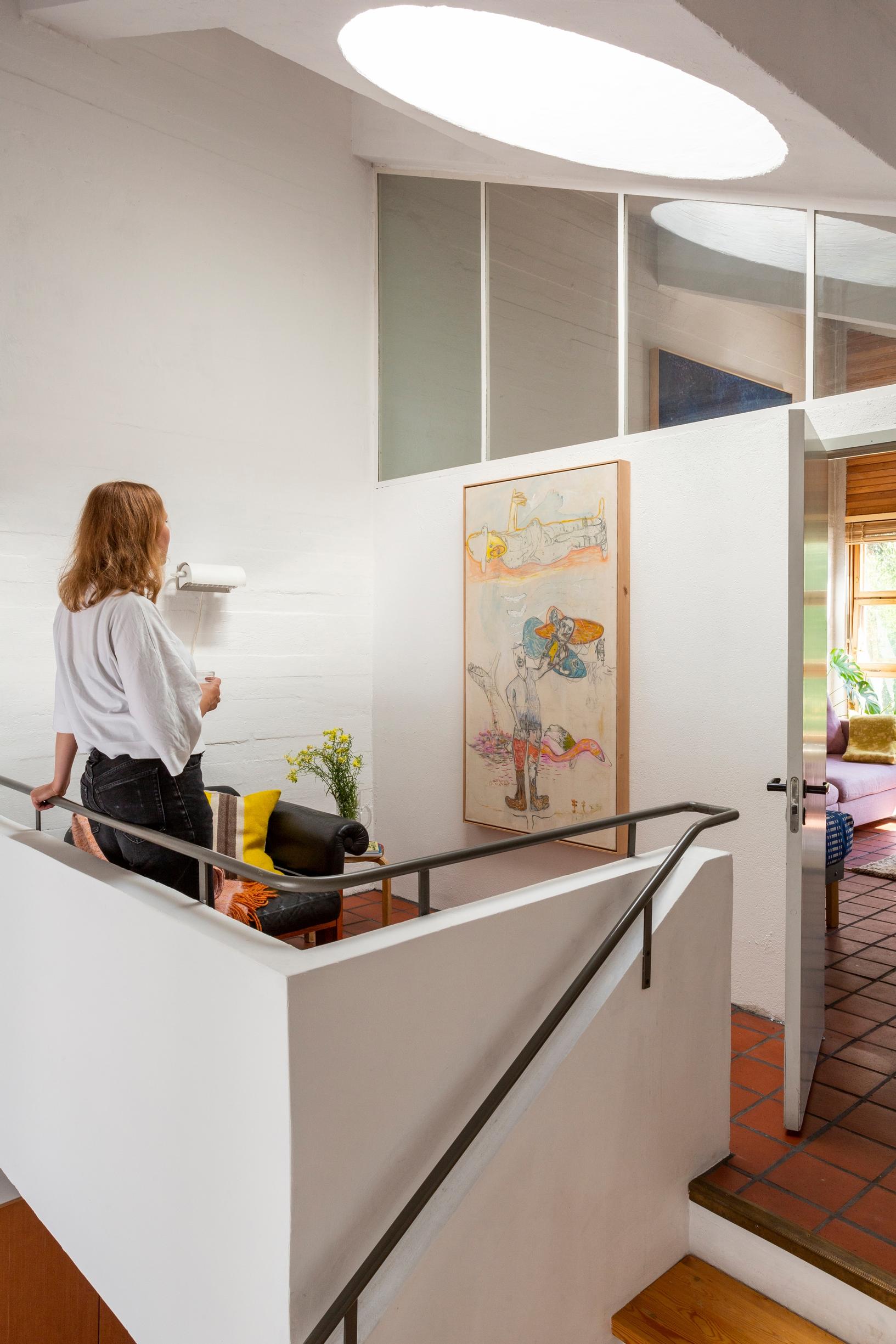 Heini-Tuuli Onnela standing, leaning on a railing and looking at the painting, light coming from the skylight