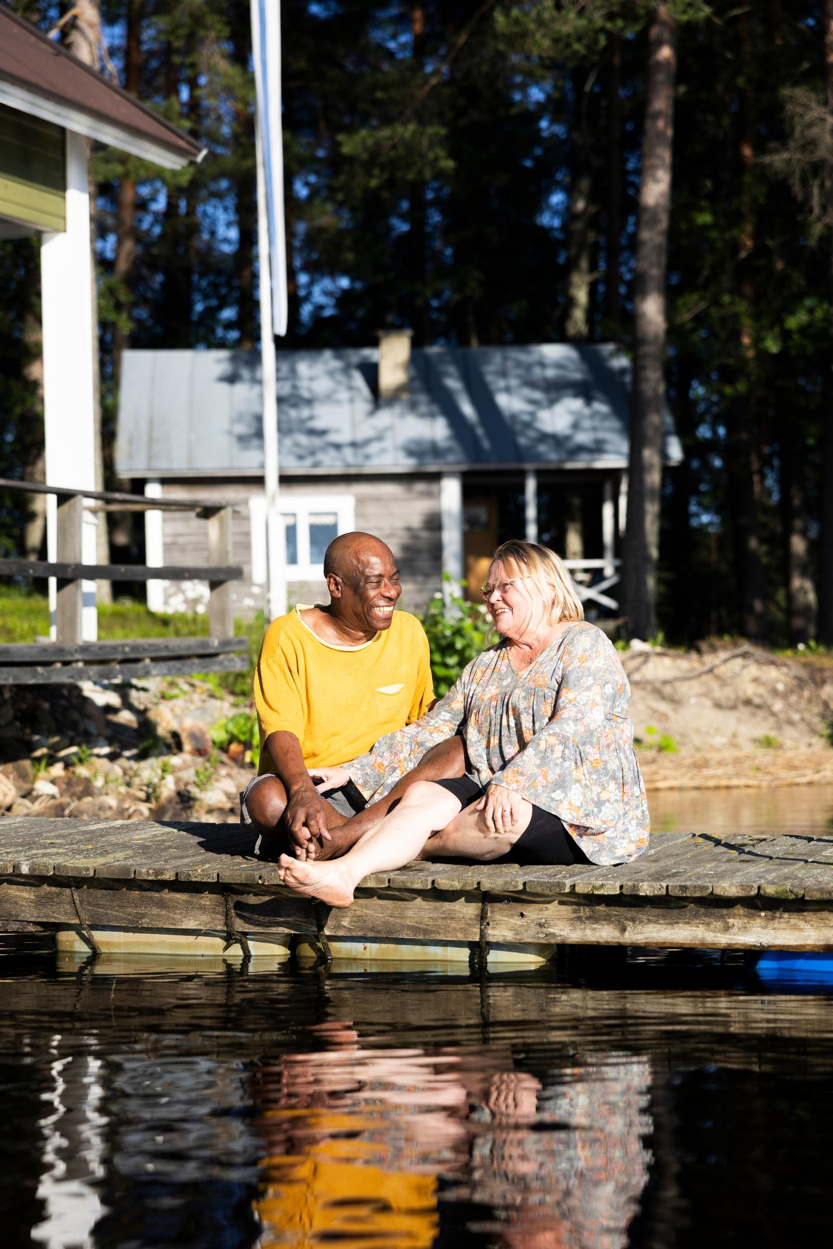 Edson and Helena sit smiling on a dock, looking at each other, with the cottage in the background