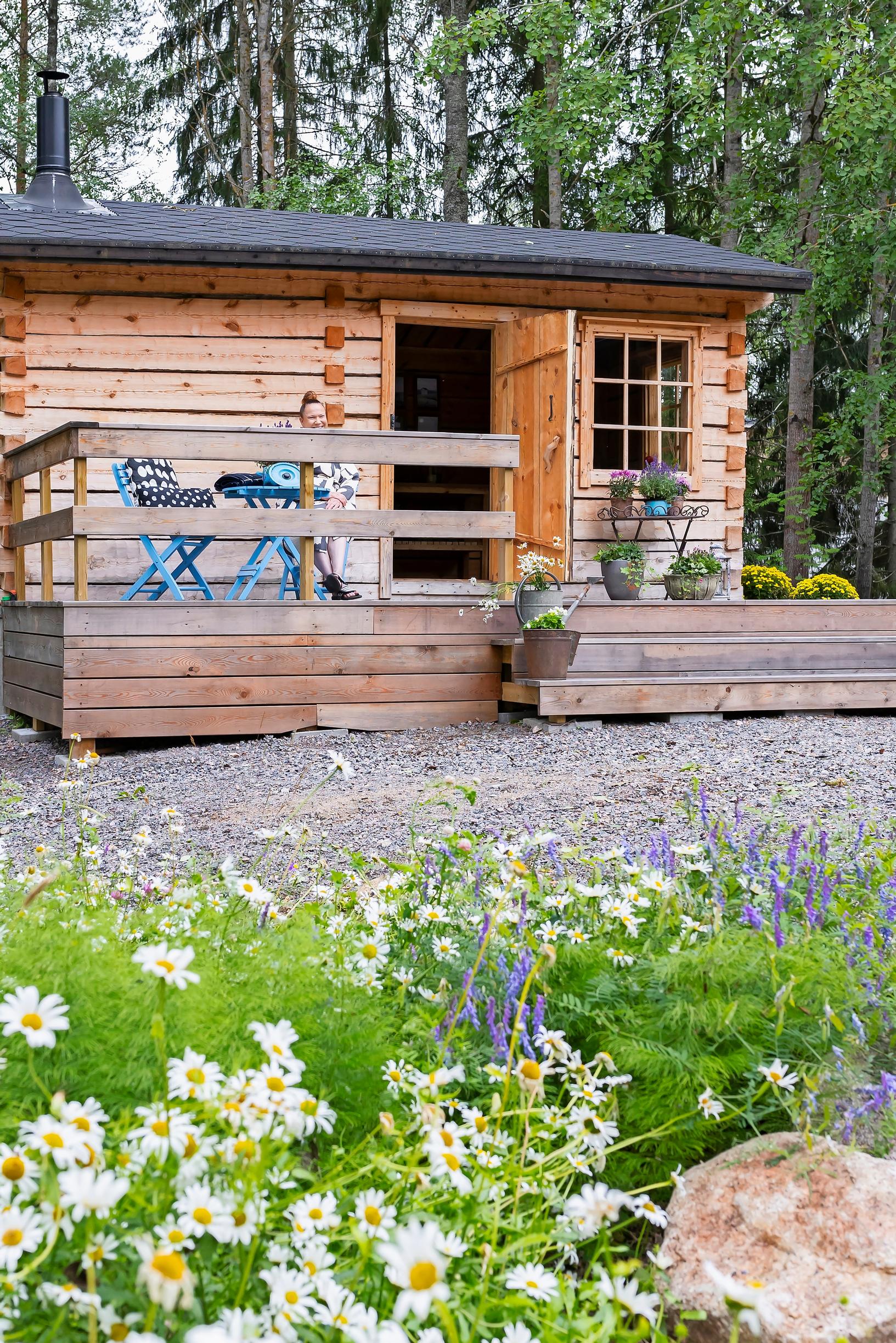 a flowering meadow in the foreground, gravel, and a sauna cabin in the background
