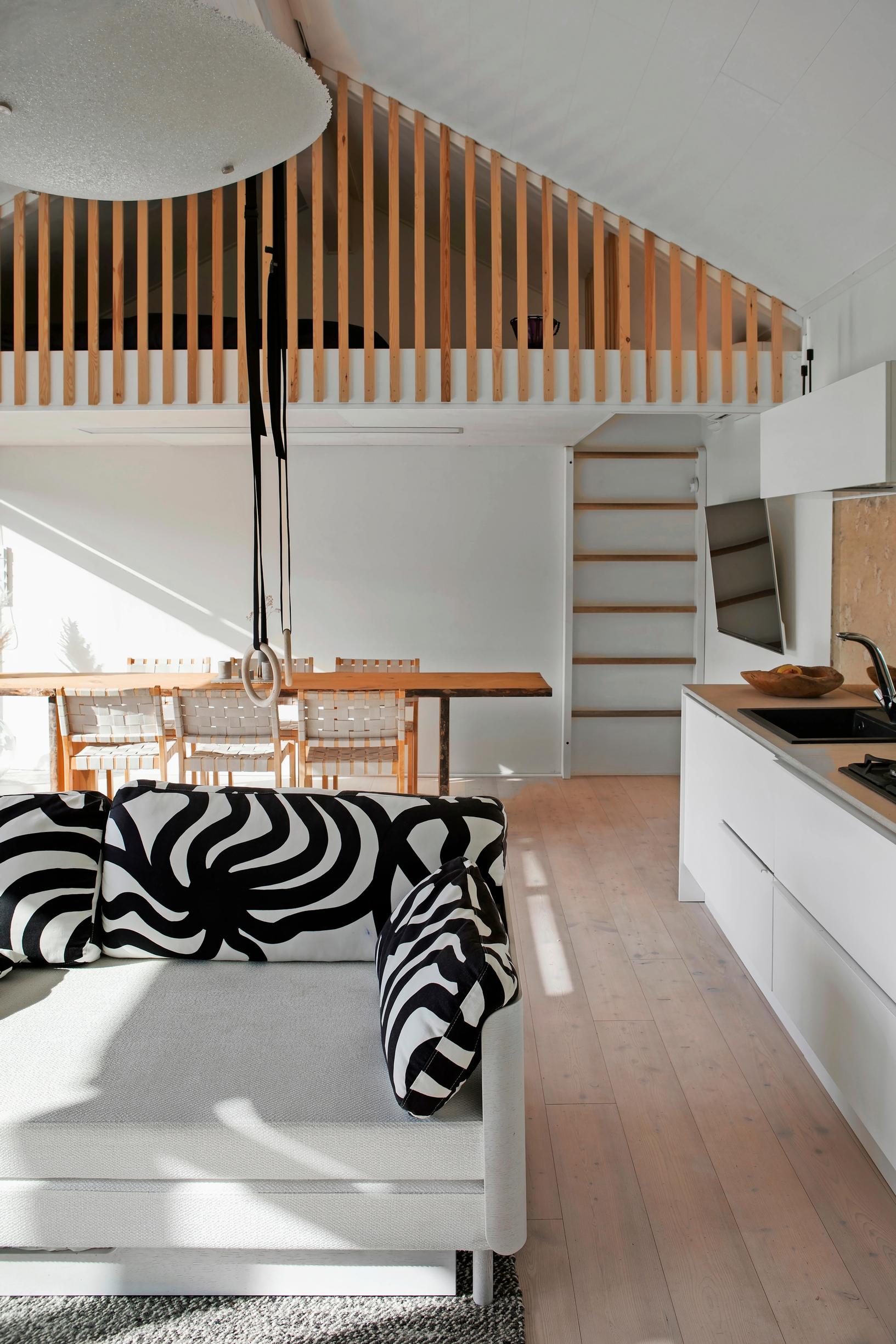 In the cabin-like space, the kitchen is on the left, a sofa in the foreground, and a dining area behind the sofa. A loft is above the dining area.