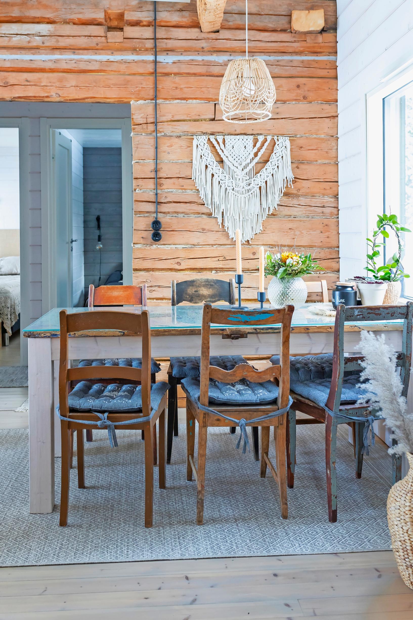 Old chairs around a large table, a wooden floor, and a log wall with a macramé piece