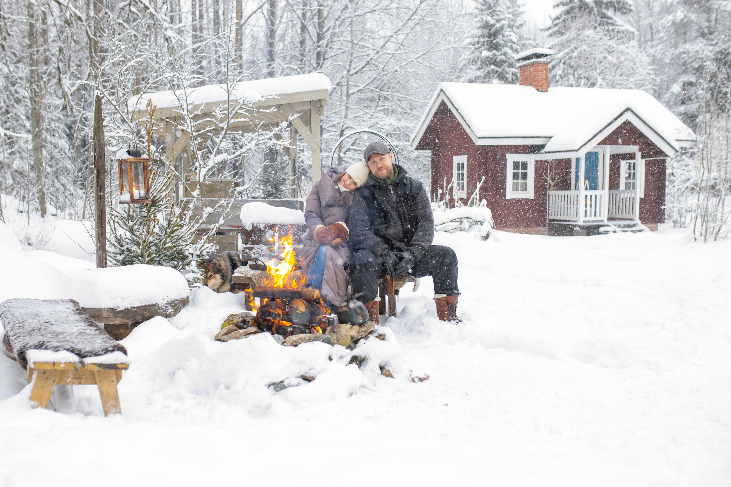 Man and woman by a campfire in winter, with a red cottage in the background.