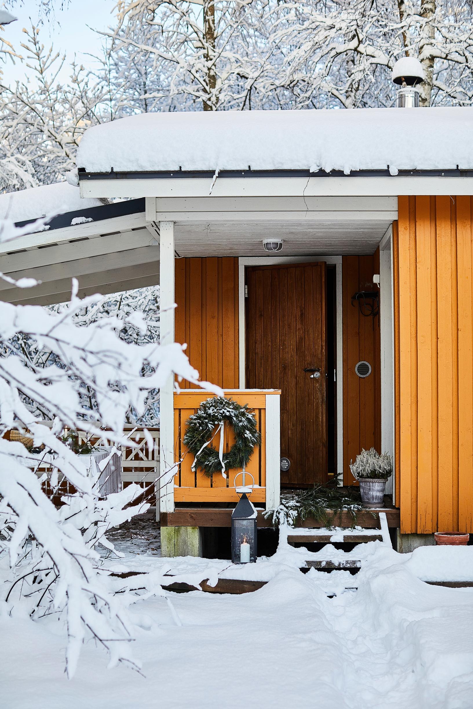 A yellow house’s front door, a wreath, and a snowy tree
