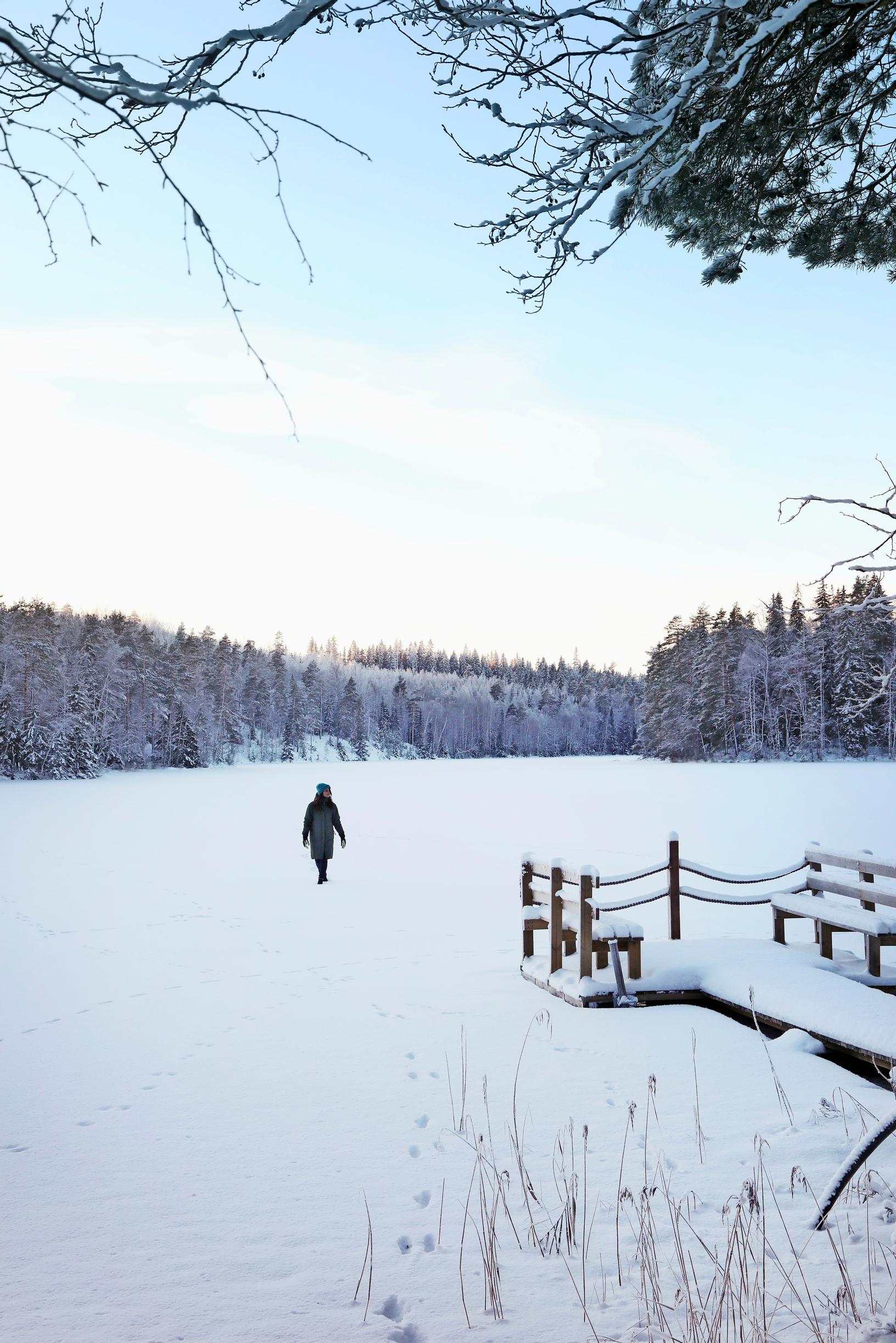 Ulla Parviainen walking across a snowy lake, with a pier and reeds in the foreground and forest on the far shoreline.