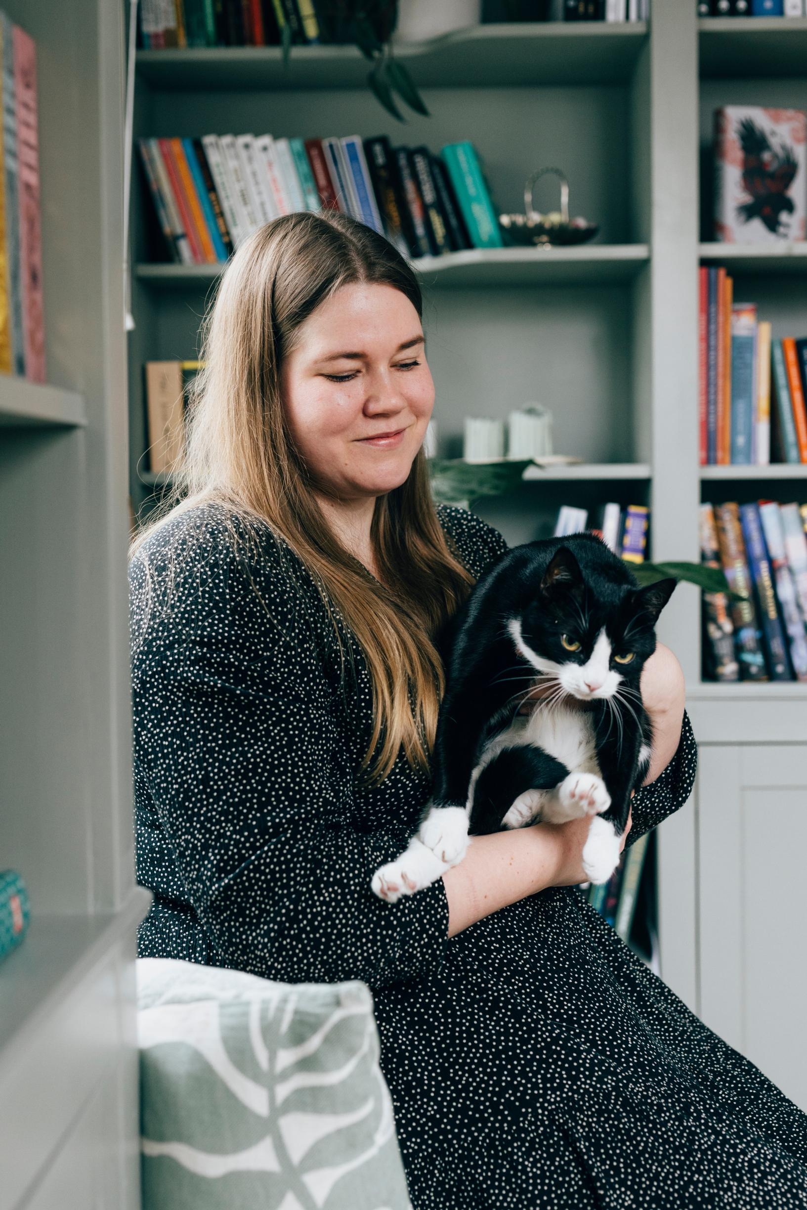 Kaisu has long dark hair and is wearing a dark-patterned dress. She is holding Helvi, a black and white cat, in her lap.