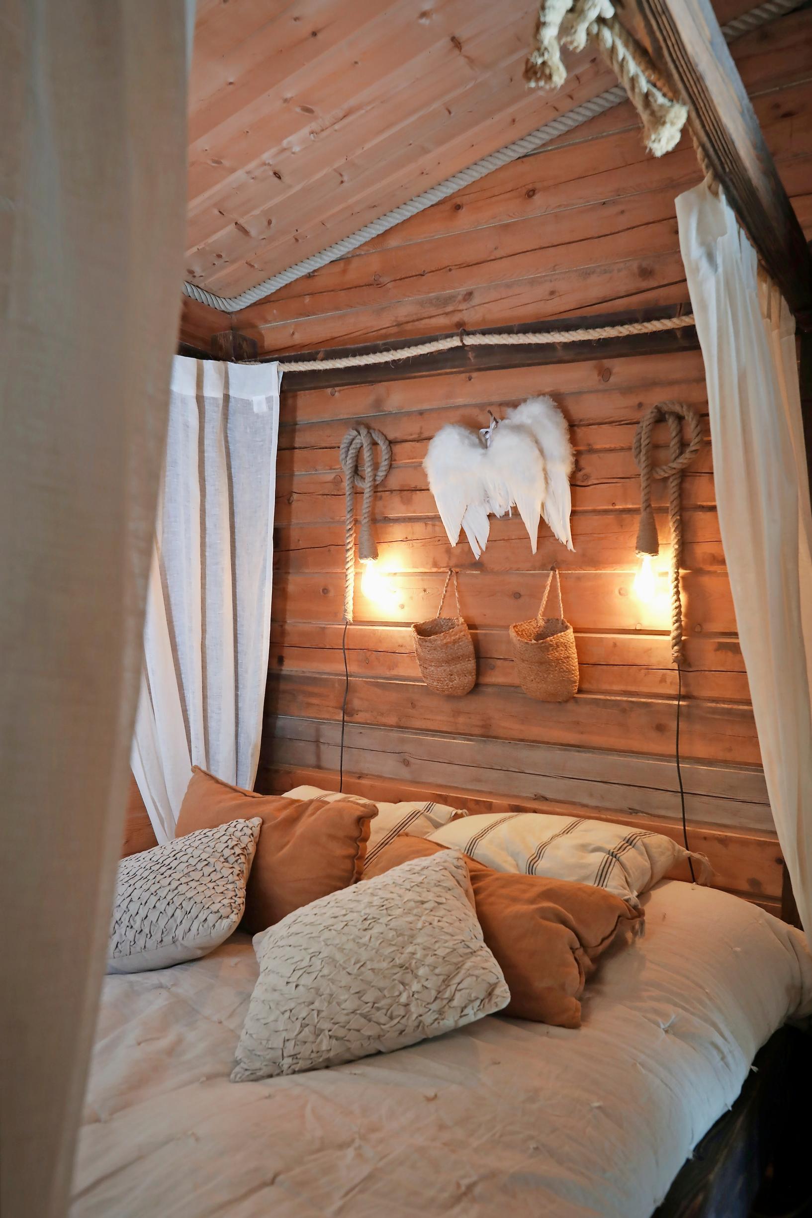 A softly-lit bed, light curtains forming a canopy, lamps on the wall, jute pouches, and angel wings.