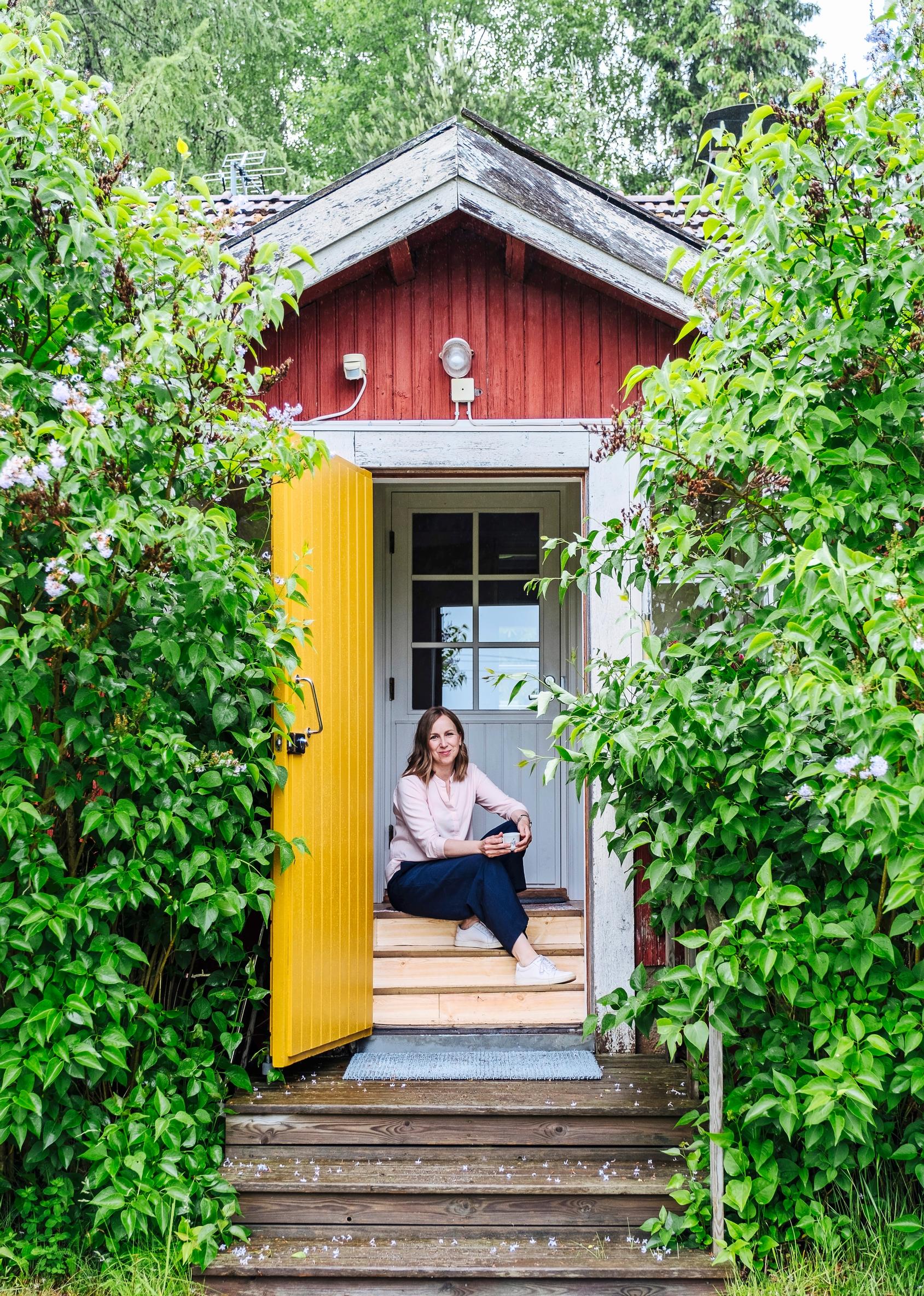woman sitting on the steps of a red wooden house with a yellow door and lilacs