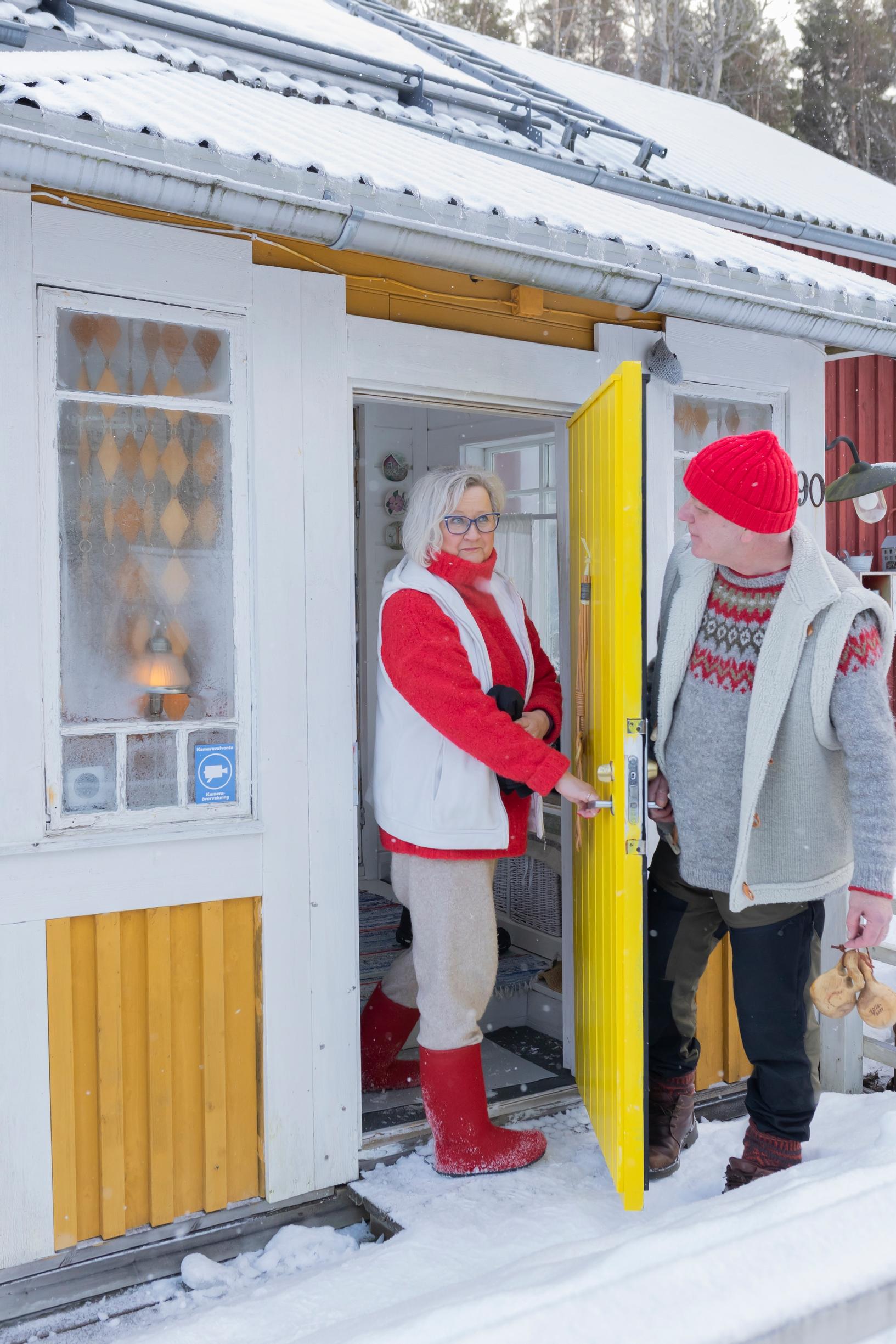 Anne and Matti standing at the door of a yellow wooden house, looking at each other