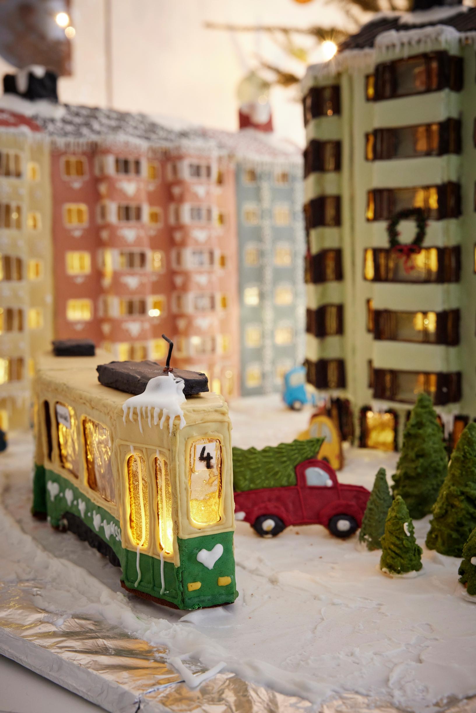 A gingerbread city block featuring a 1950s apartment building with bay windows covered in green and pink royal icing. A streetcar runs through the middle of the block.