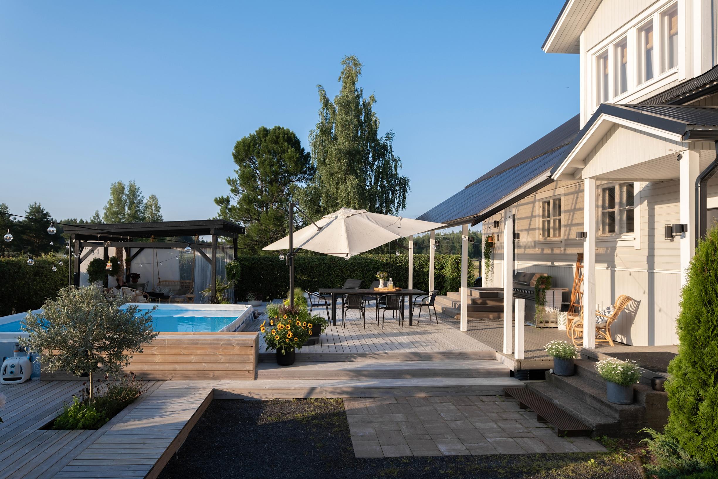 A large deck, a swimming pool, and a pergola in front of the light-colored wooden house.