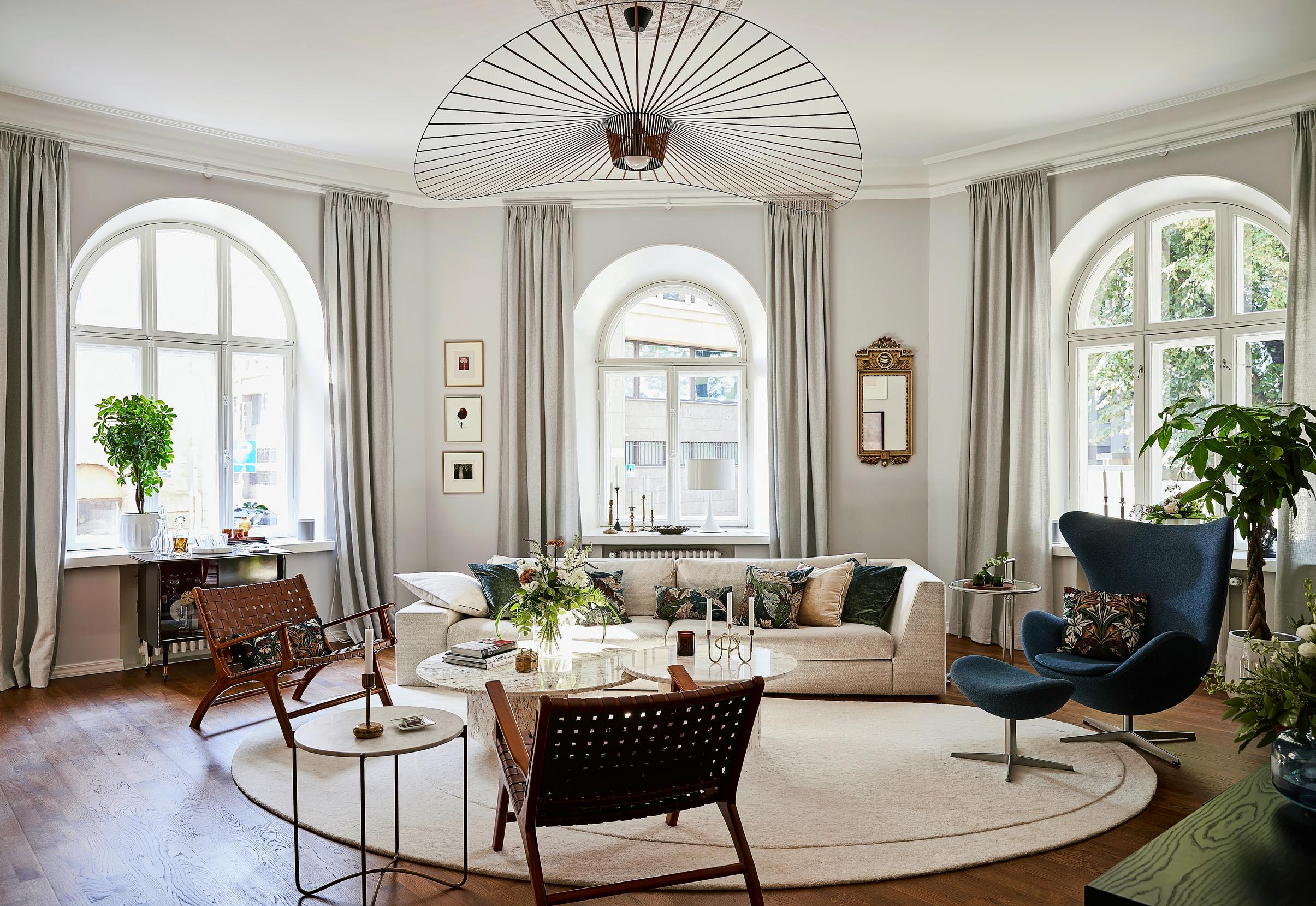 In the angled living room with arched windows: a light-colored sofa, lounge chairs, and a petrol-blue Egg chair