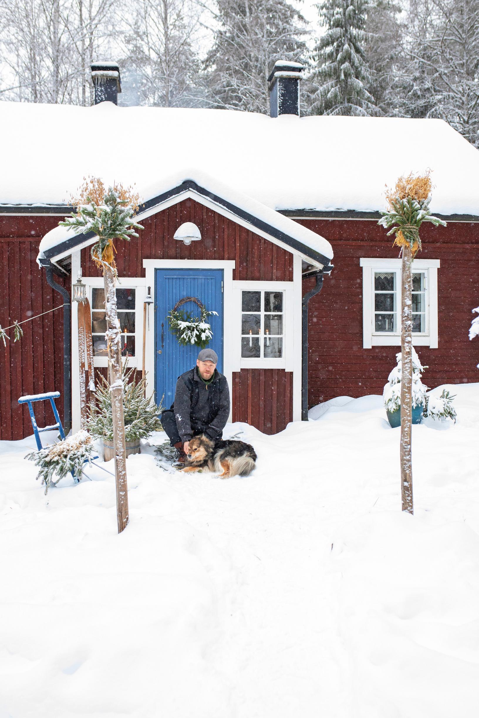 A man and a dog in front of a red cottage in winter.