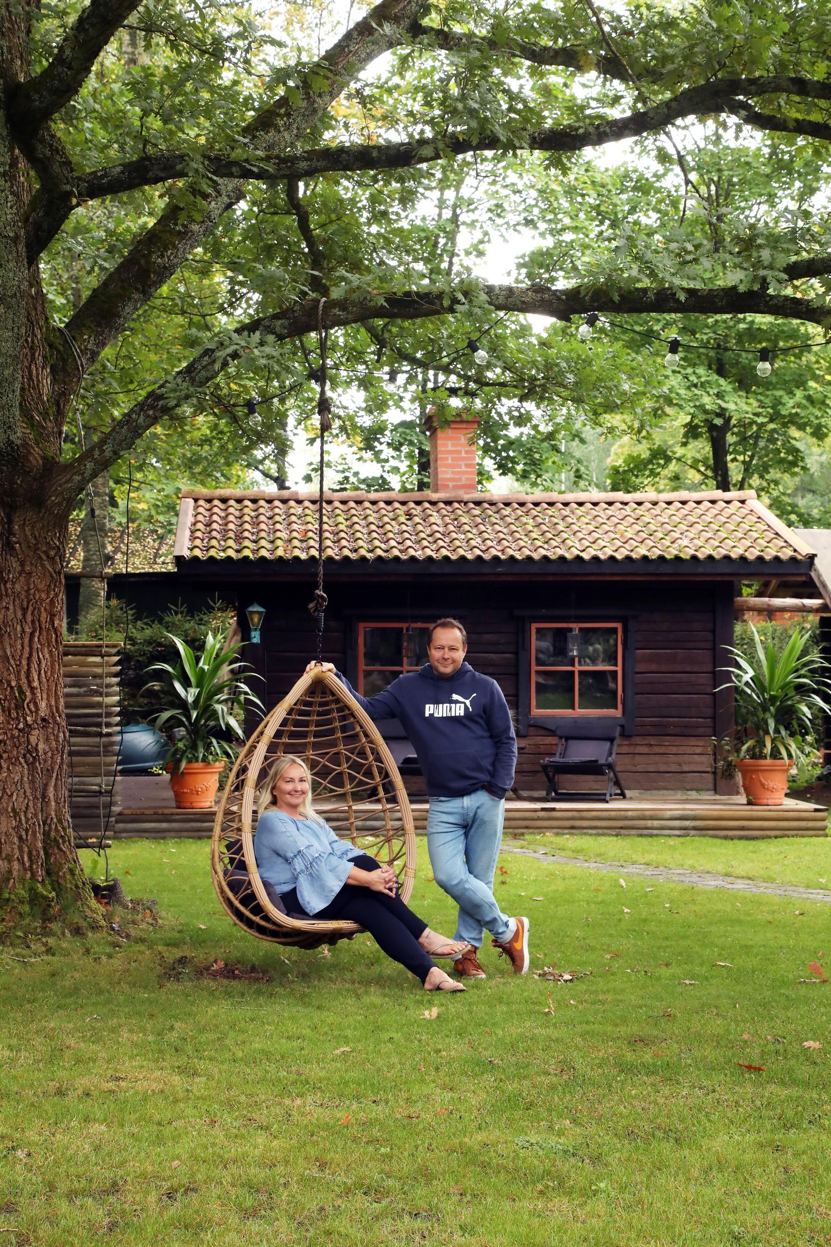 Katja sits on a hanging rattan chair, while Kimmo stands beside her. A wooden sauna building is behind them.