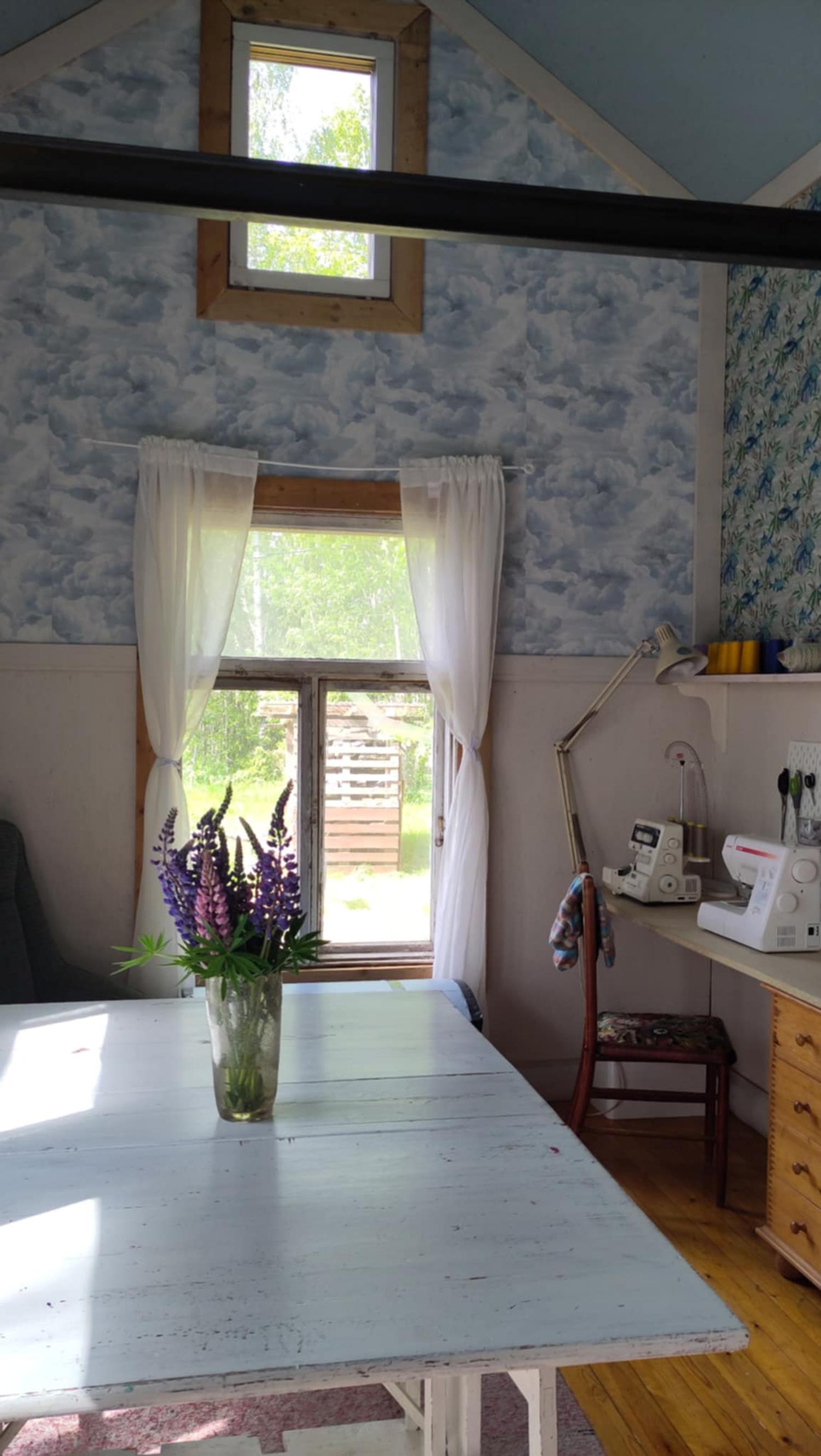 The room features a light-painted wooden kitchen table, patterned blue wallpaper, and white curtains at the windows.