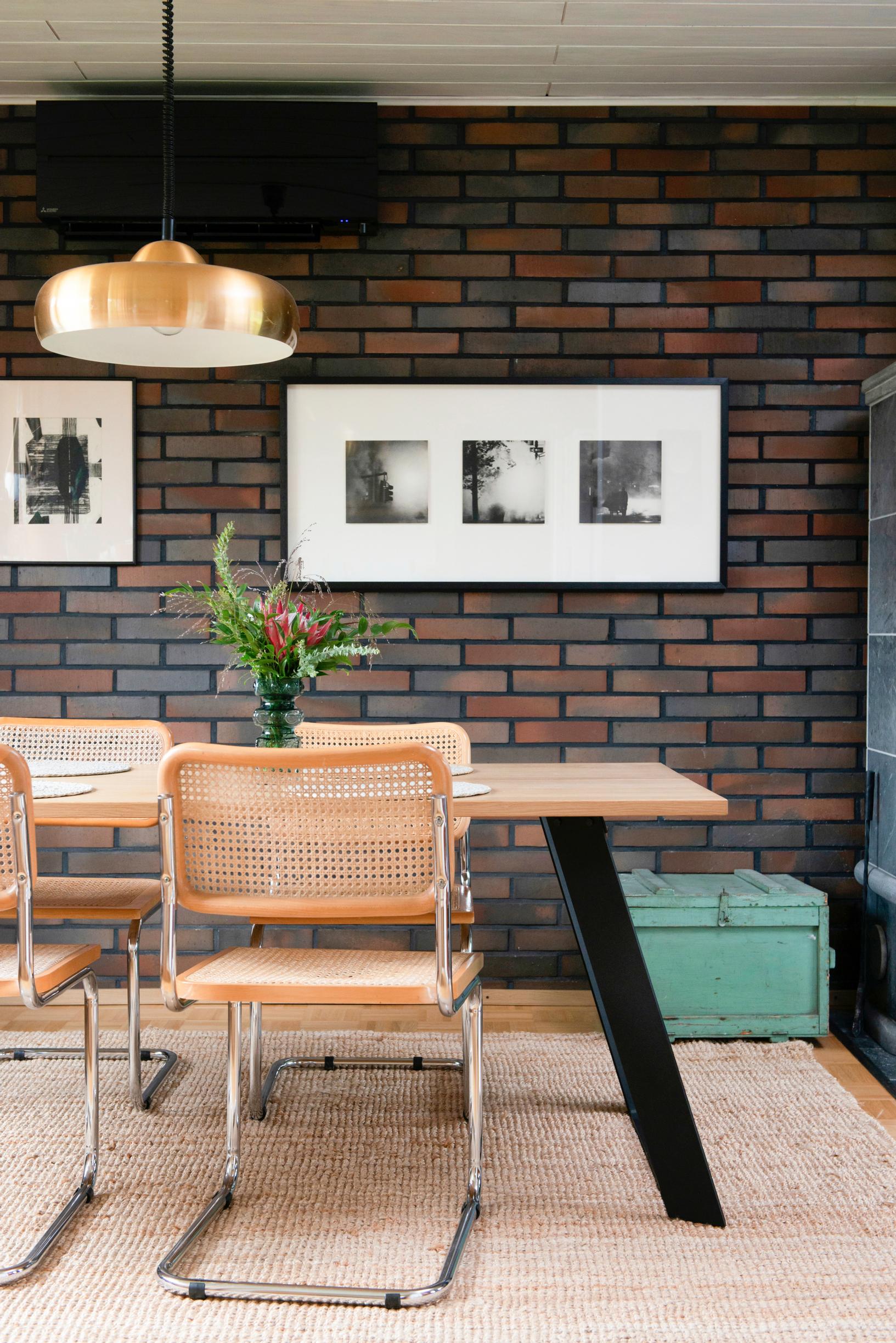 Two pieces of art hang on the variegated brick wall. In the foreground, there is a dining table with chairs.