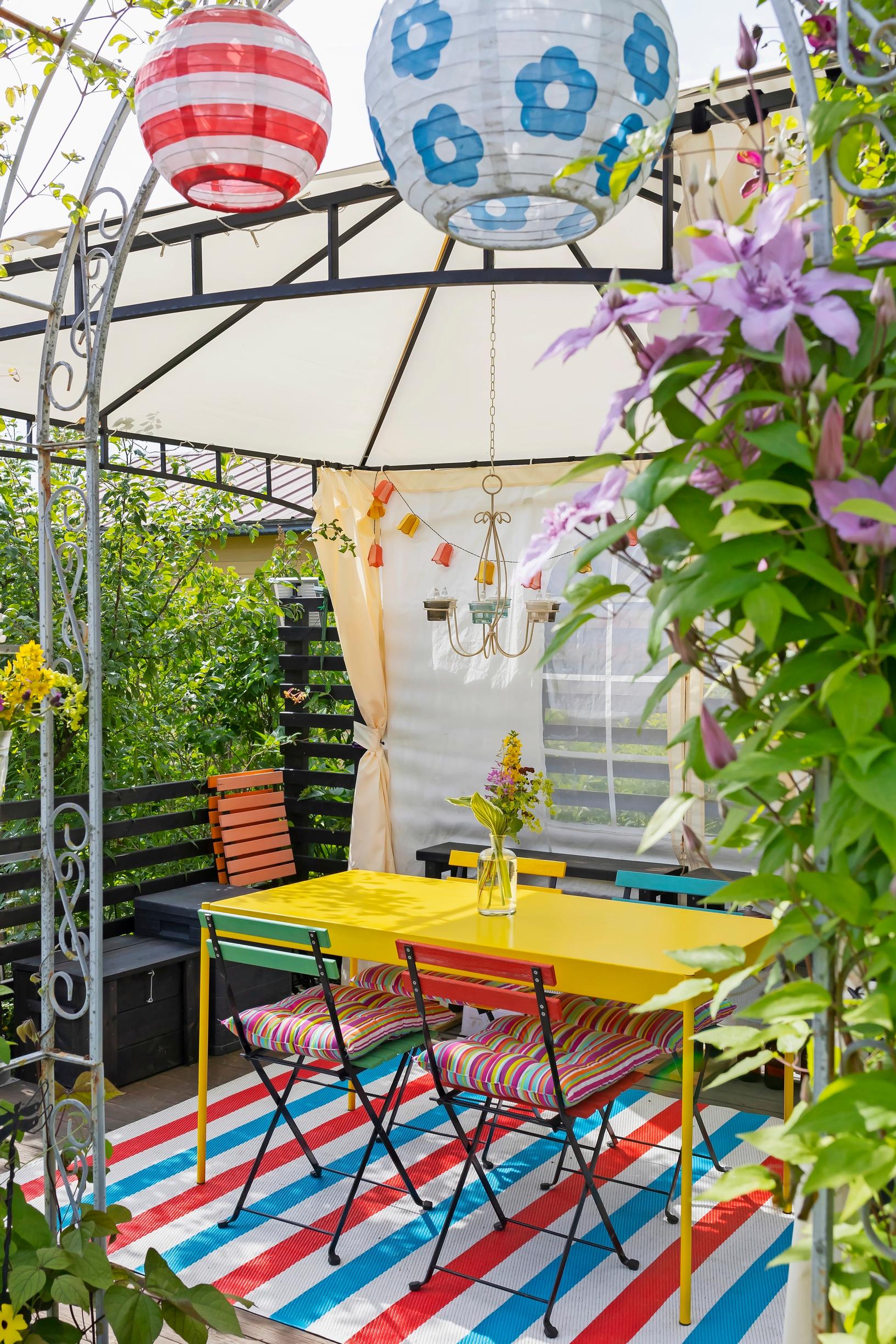 a yellow table and colorful chairs under a canopy, a striped rug, and clematis plants