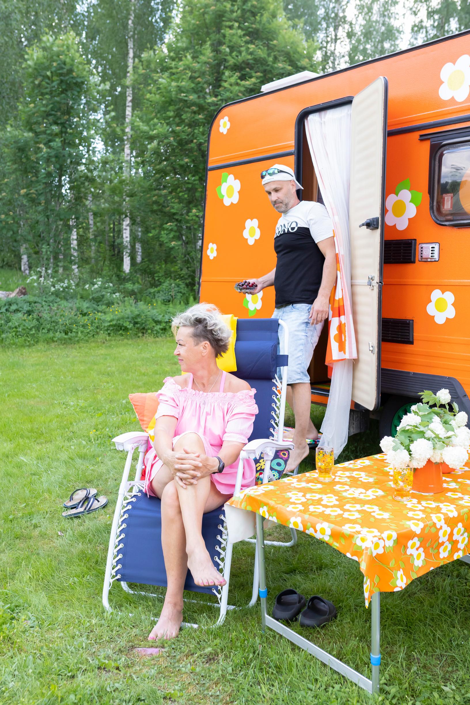 Kirsi Niiranen sits in a lawn chair in front of the colorful travel trailer, while Jaakko Timonen steps out of it