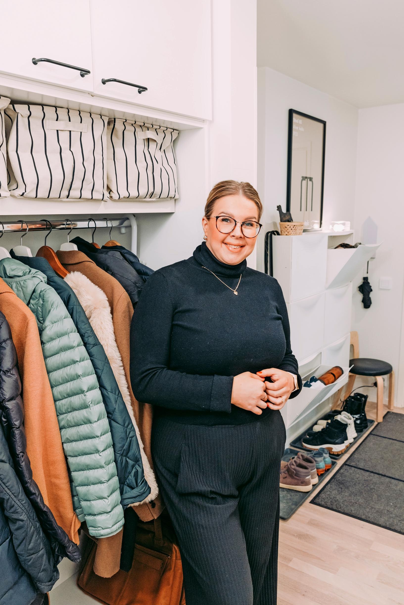 A woman stands in the entryway in front of a coat rack.