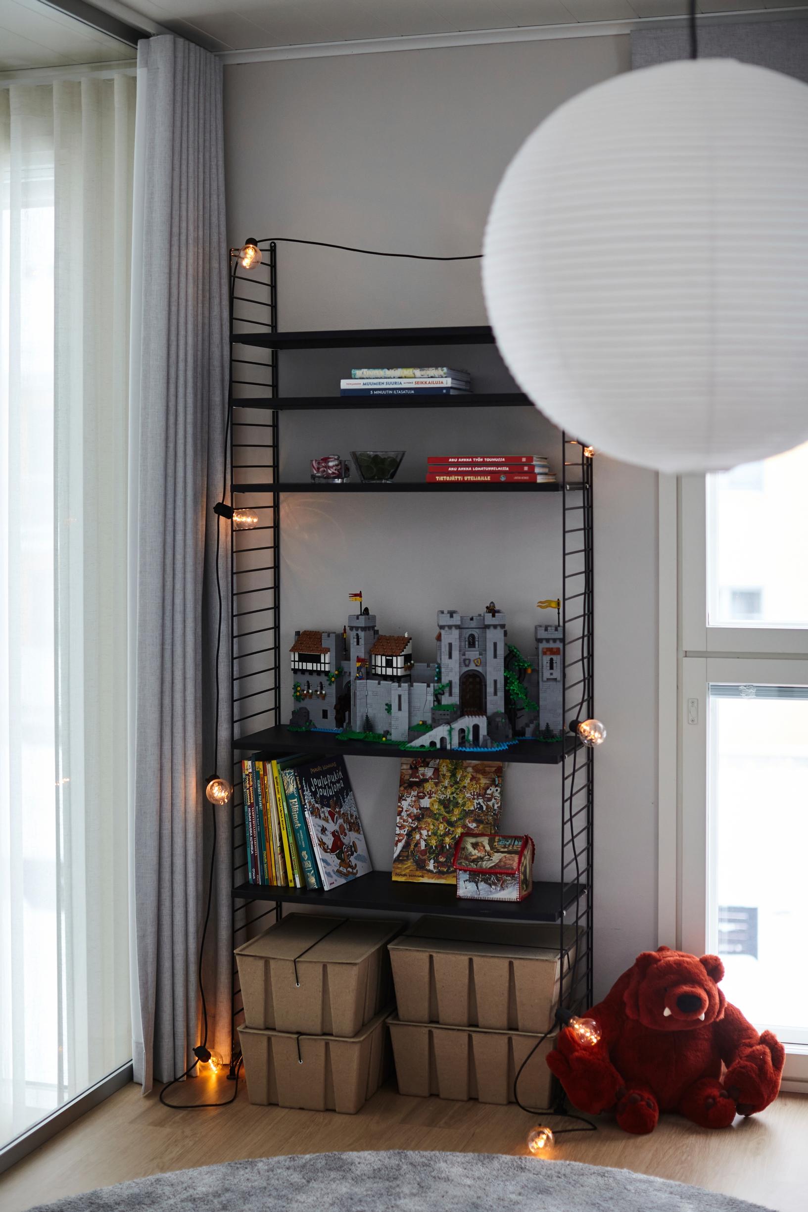 a black metal shelf, a Christmas light string, a plush toy, and a rice paper lampshade in the children’s room