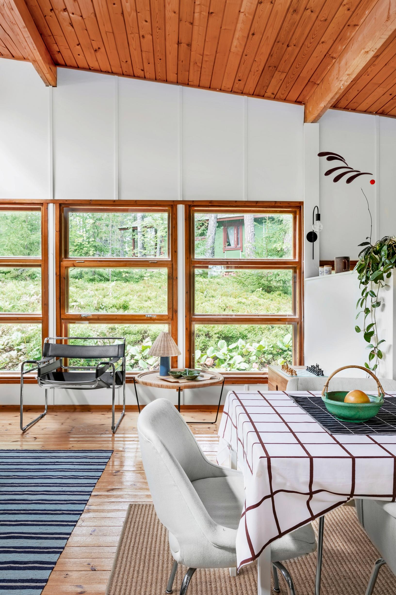 A large paned window overlooking nature, an armchair and a coffee table, and a dining set on a wooden floor.