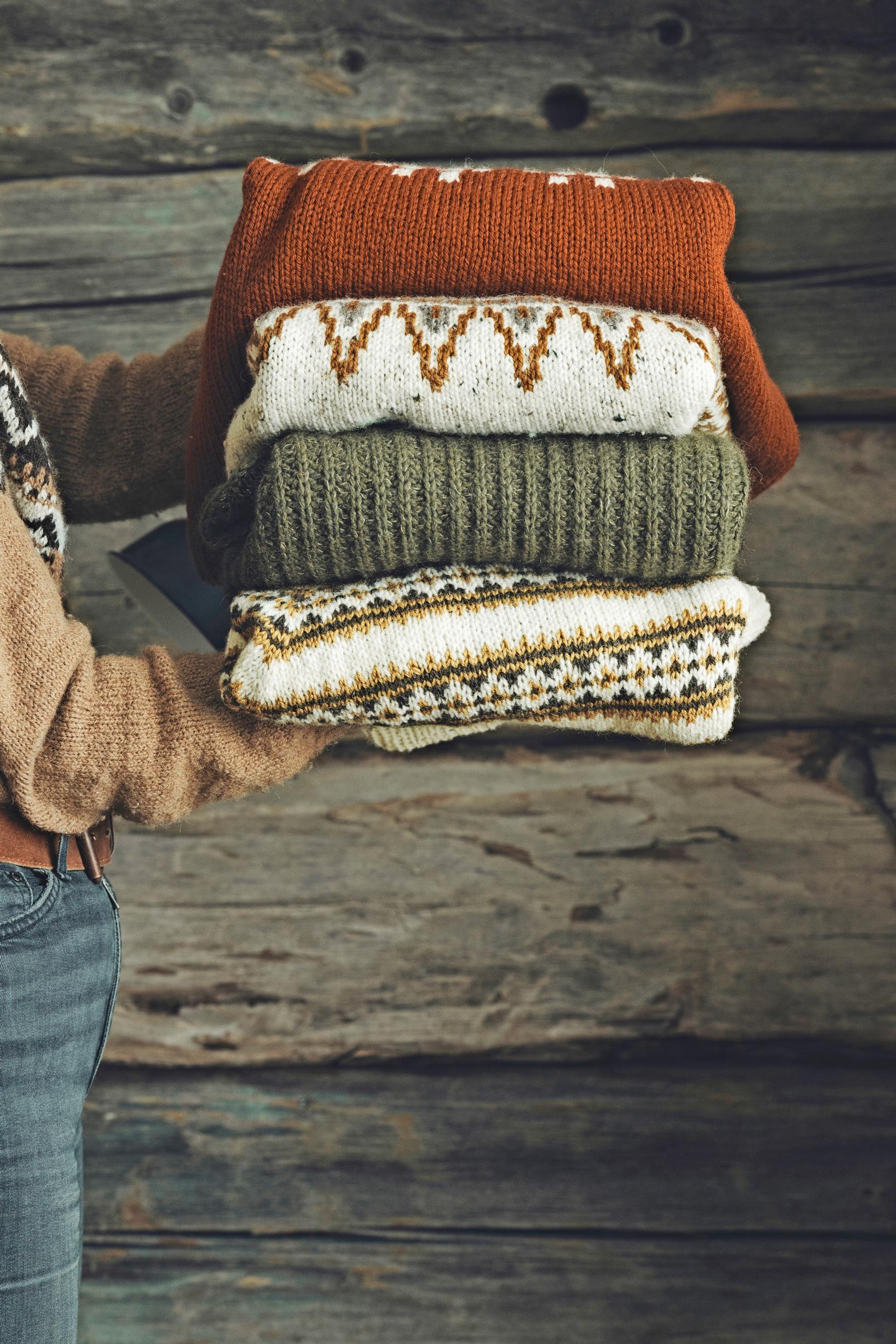 A stack of knits in her hands, with an old log wall in the background.