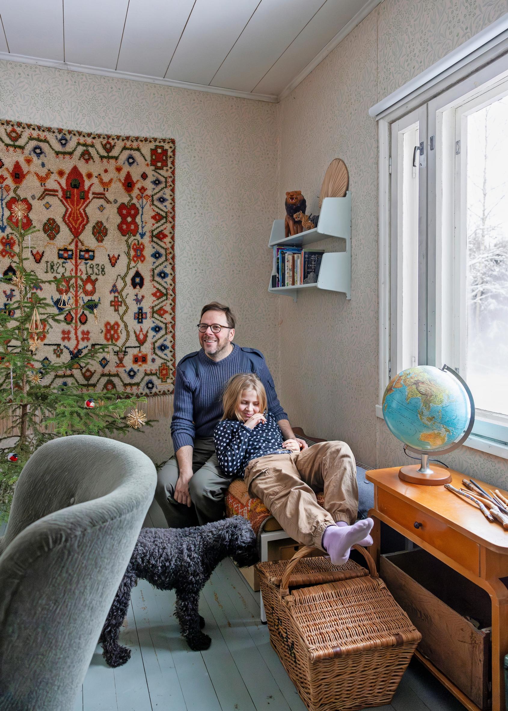 Daughter Elsa and father Klaus Aalto on the sofa at the cottage in Kisko, with a ryijy in the background, dog beside them, and a globe on the windowsill