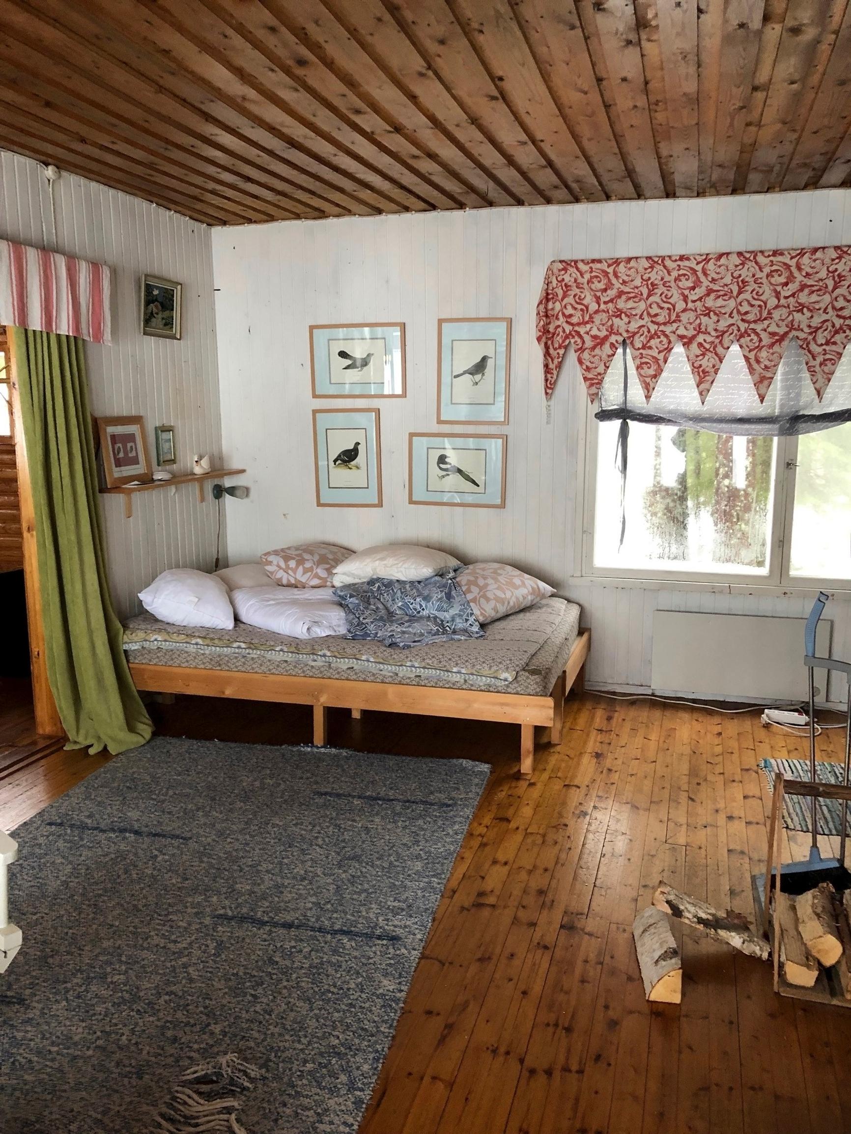 A double bed with lots of pillows, bird paintings, wooden floor, a rug, and red curtains