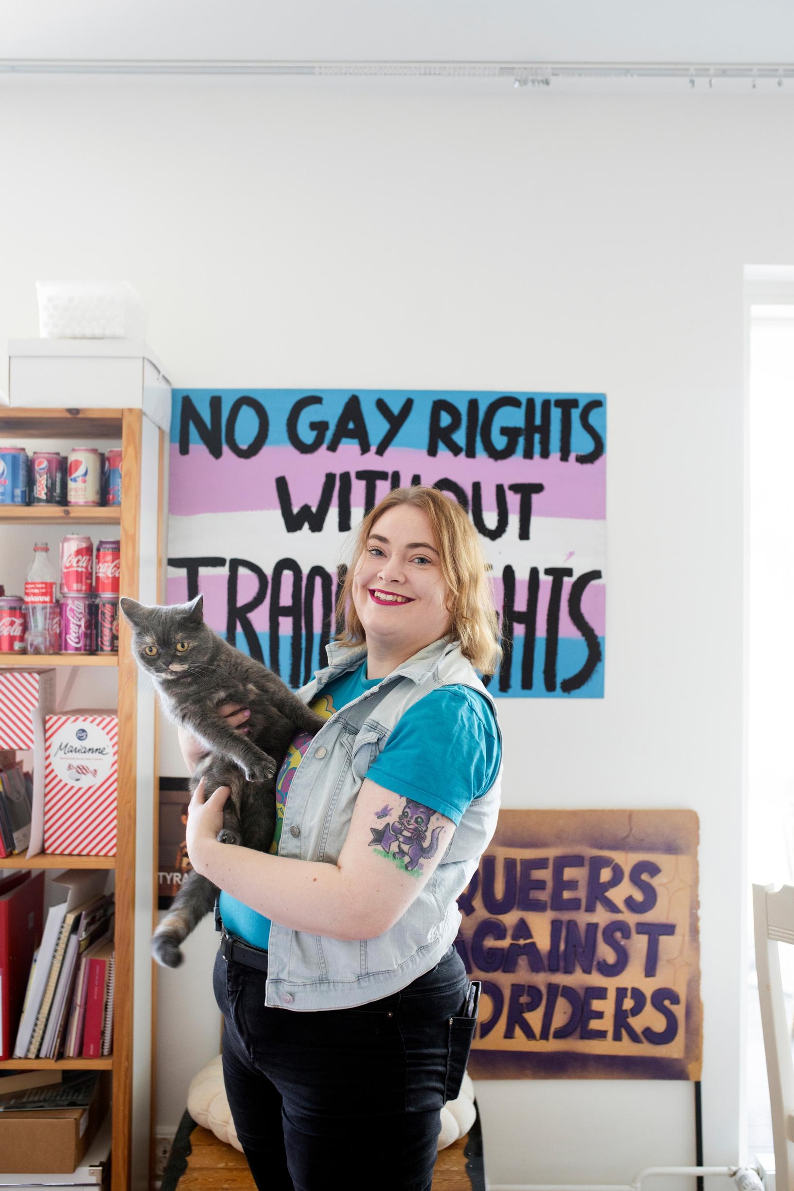 A woman holds a cat in her arms. The posters in the background read ‘No gay rights without trans rights’ and ‘Queers against orders’.