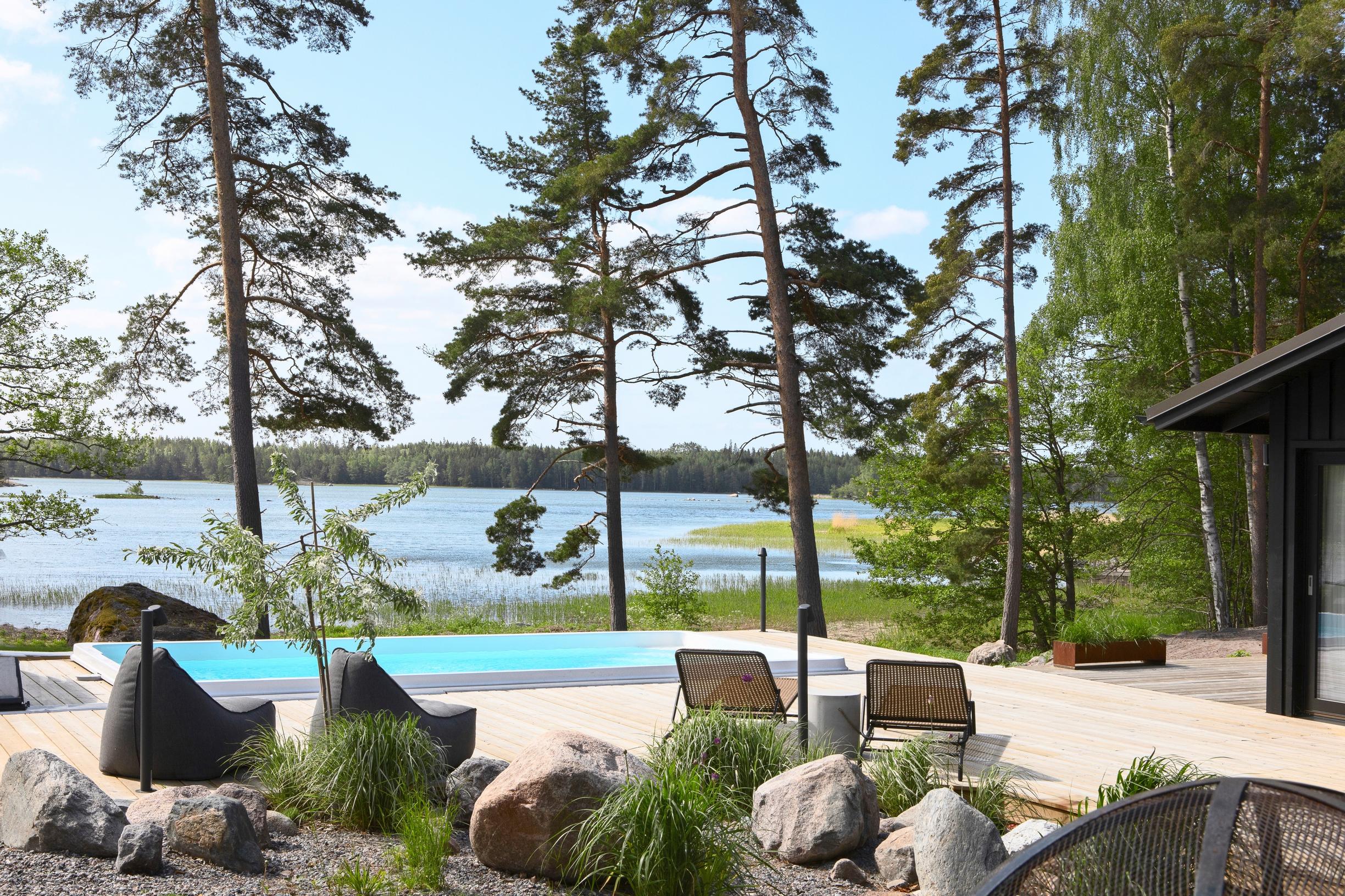 On the terrace of a summer home sits a swimming pool, with views of the sea and trees, and garden furniture in the foreground