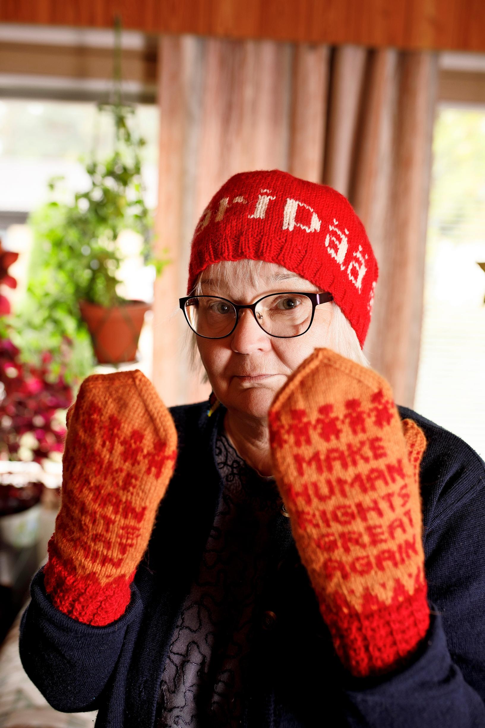 A woman looks at the camera and shows mittens that read ‘Make human rights great again.’ She’s wearing a red hat that says ‘extremist.’