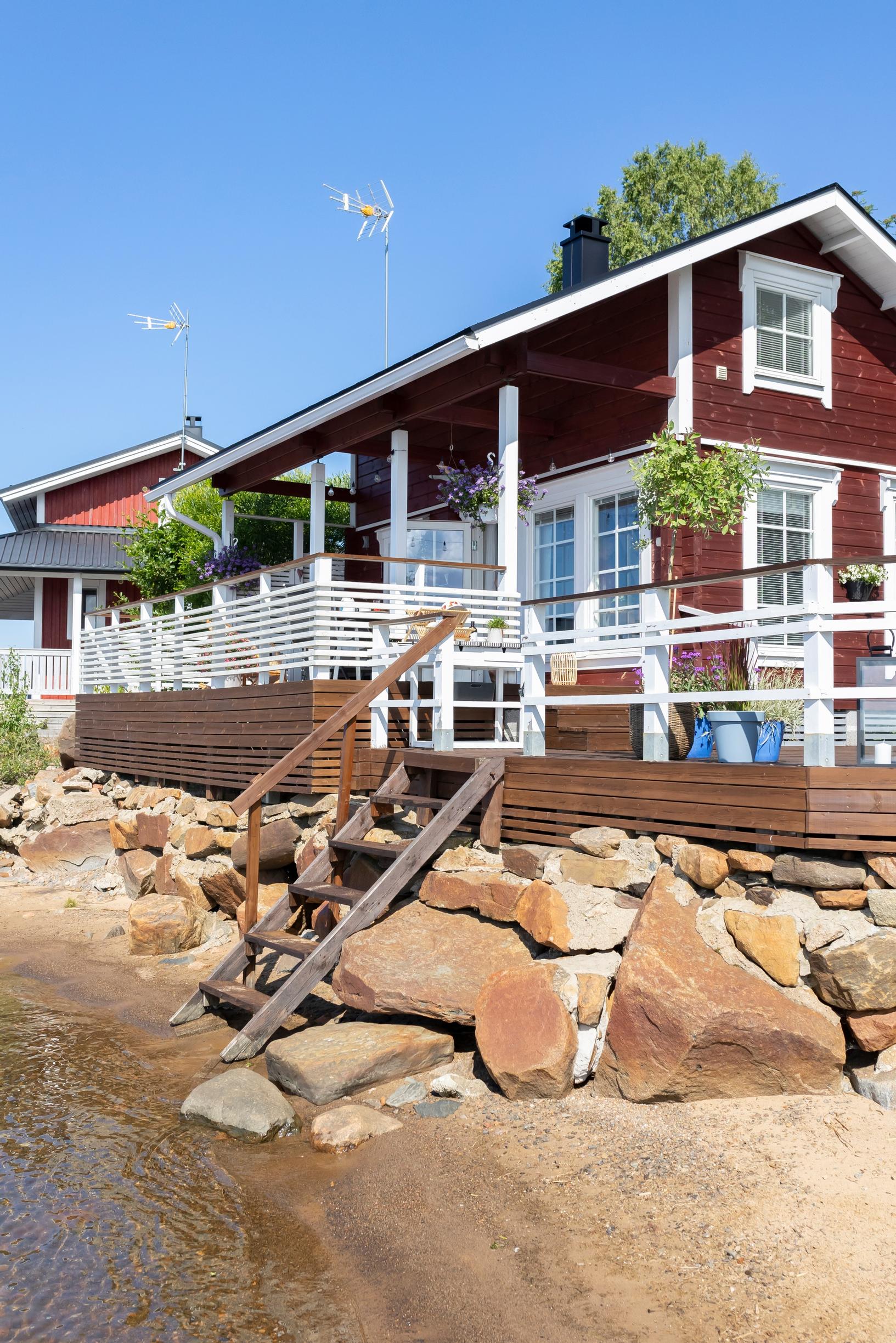 a red cottage and a terrace by the shore, with steps down from the cottage, a sandy beach, and stones