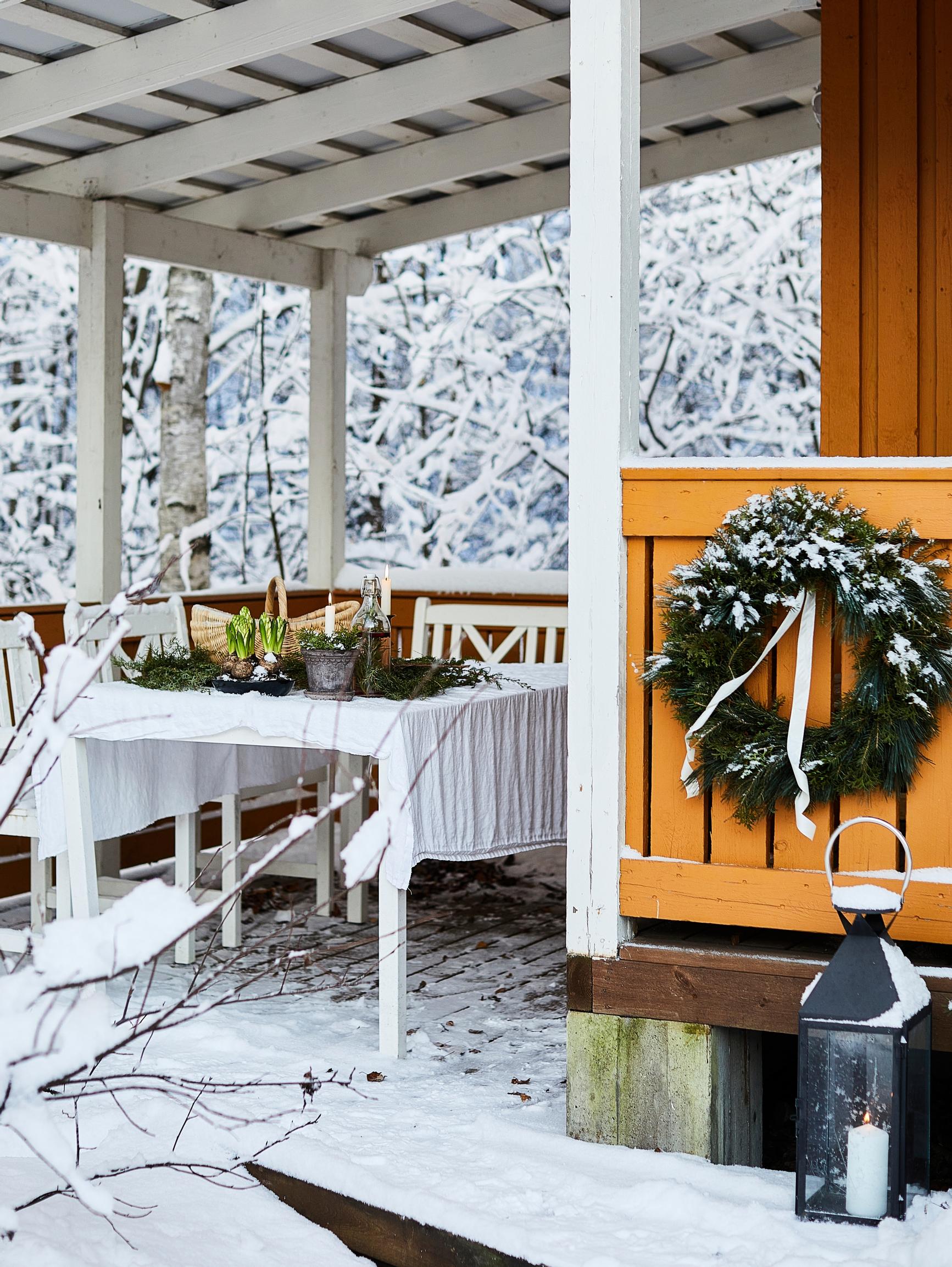 A set table on an outdoor terrace in winter, an evergreen wreath, a lantern, and snowy woodland