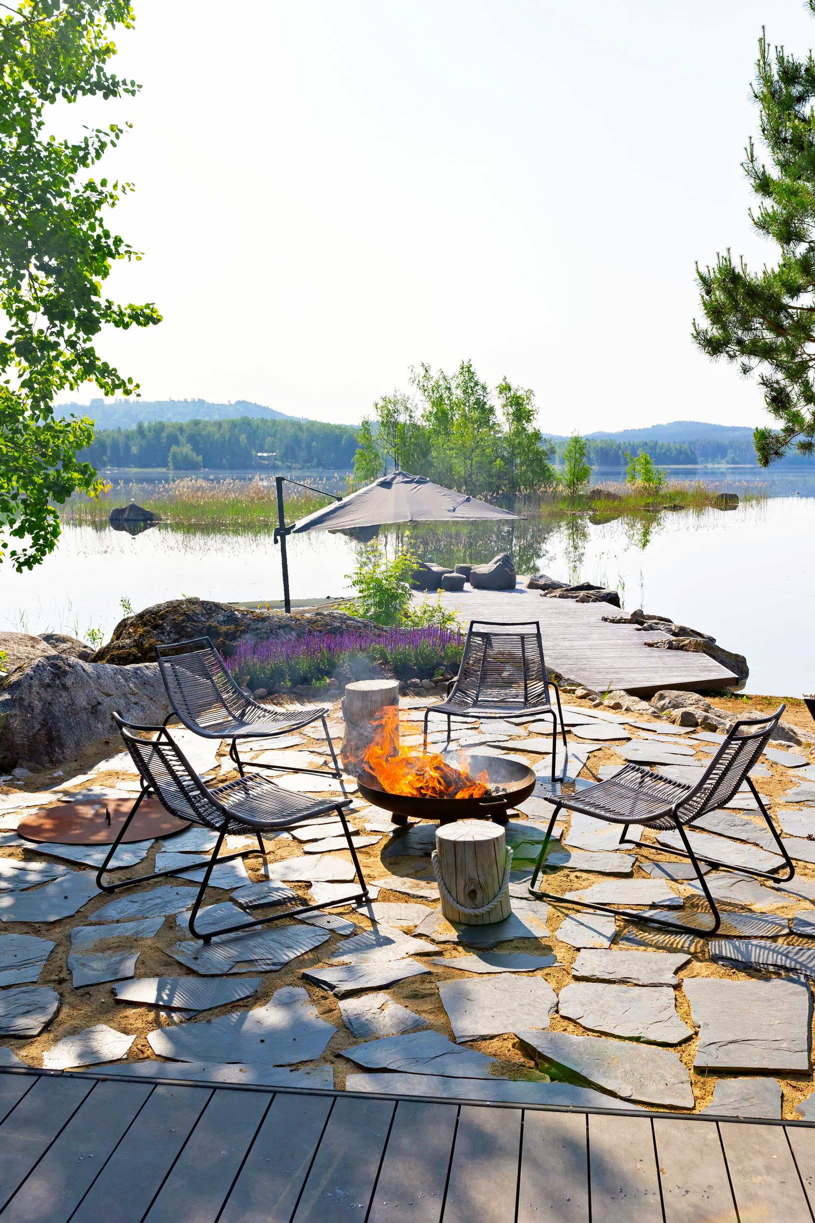 A fire in the hearth on top of slate stones, surrounded by chairs and logs, with a dock and a lake view in the background.