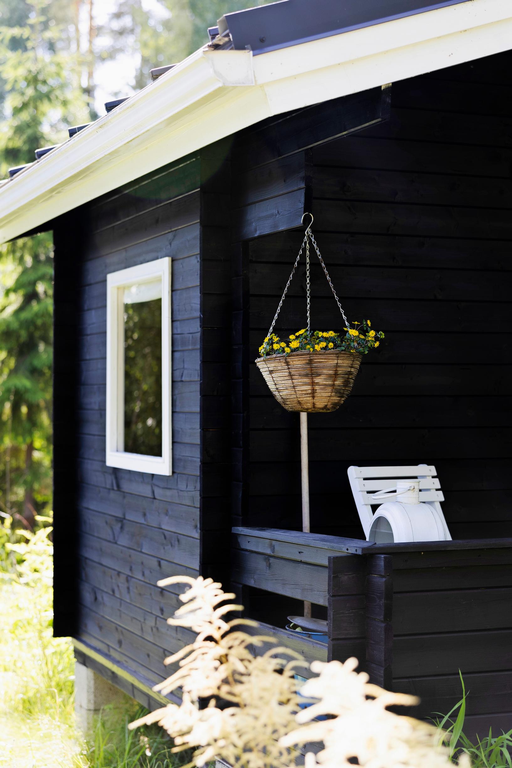 a small black log cabin with white window frames, a hanging flower basket, and a garden