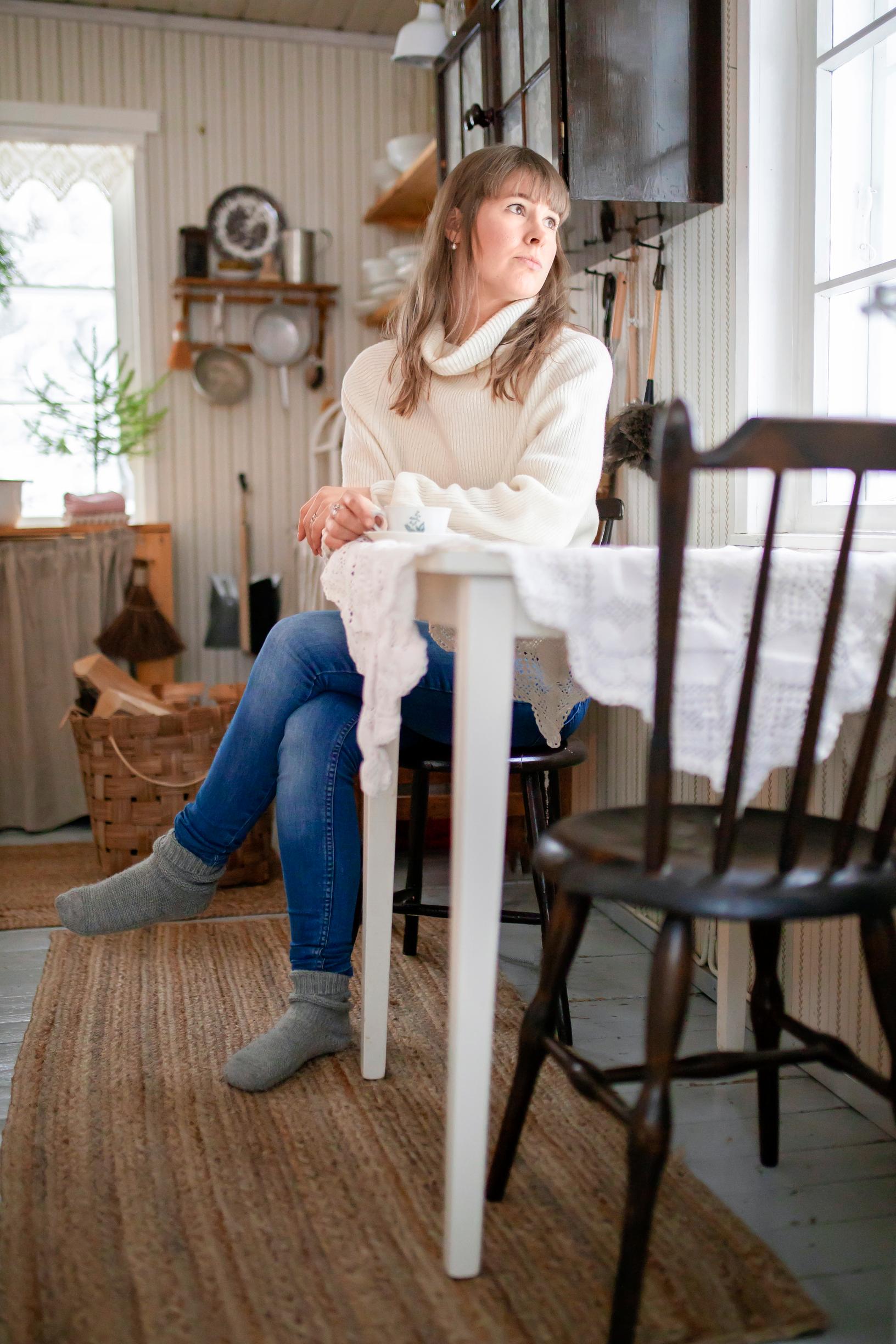 A woman looking out the window while seated at a table with a lace cloth.