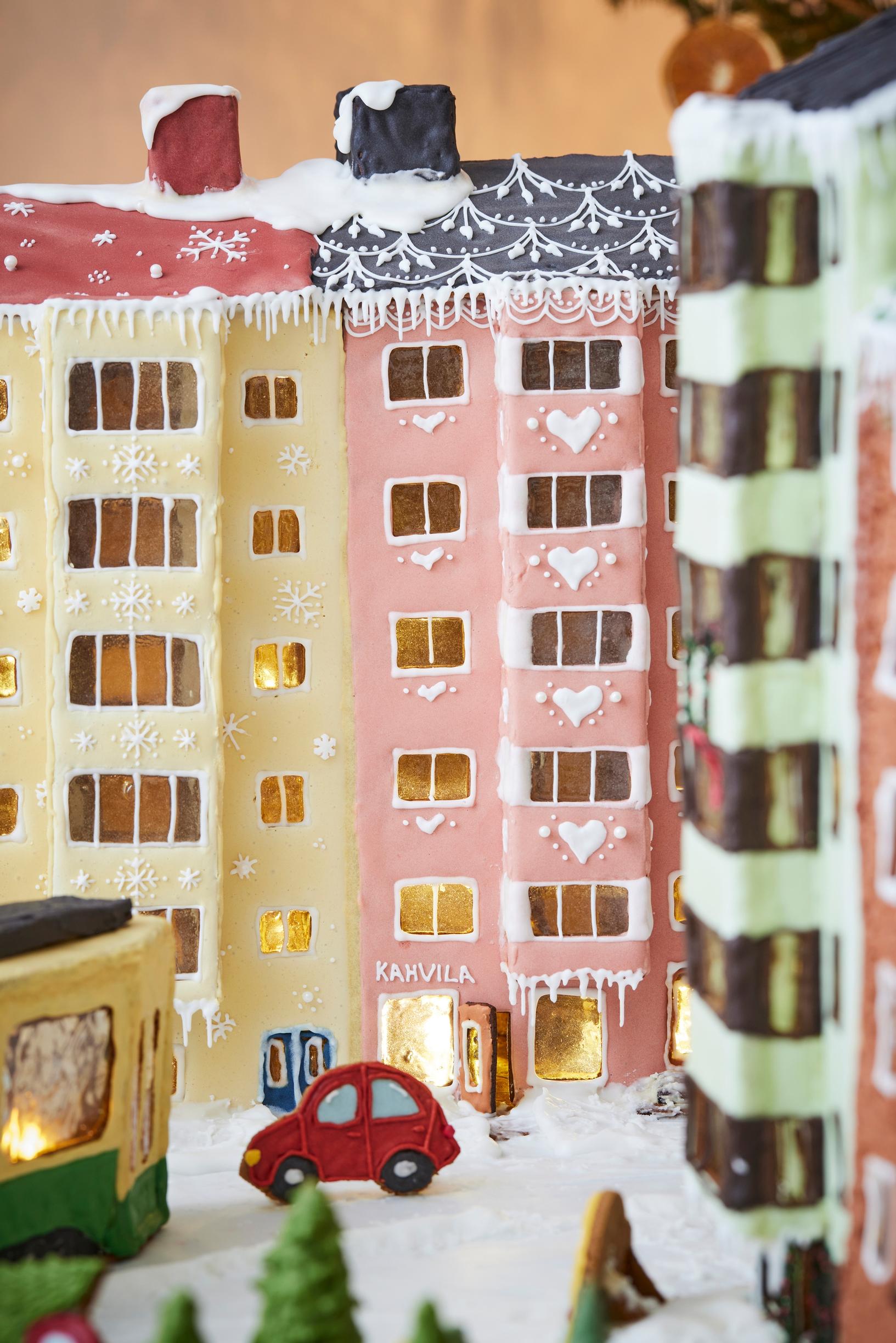 A gingerbread city block featuring a 1950s apartment building with bay windows covered in yellow and pink royal icing. A red car drives in front of the buildings.