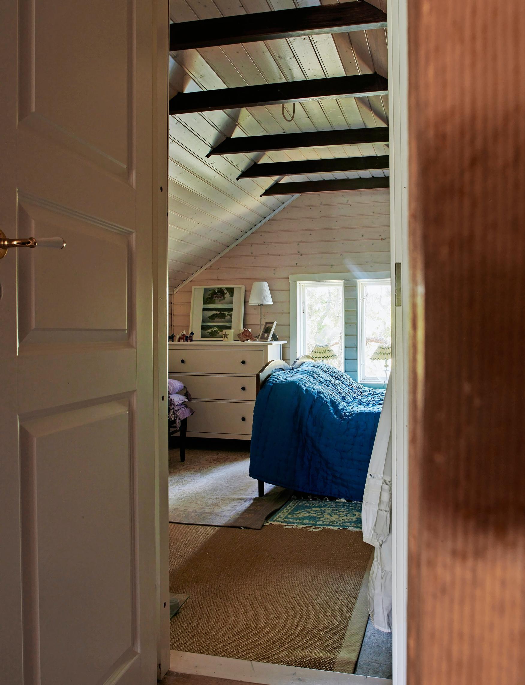 a blue bedspread on the bed, a white chest of drawers, slanted ceiling and a mirrored door